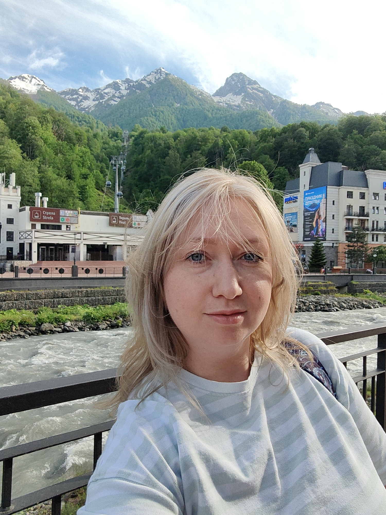 This media file features a selfie of a woman set against a picturesque mountain resort in Estosadok, Russia. In the foreground, a woman with long, slightly windswept blonde hair and blue eyes looks directly at the camera with a calm, pleasant expression and a subtle smile. She is wearing a light-colored, possibly mint green, striped long-sleeved top, with a patterned strap visible over her right shoulder.
Immediately behind her is a black metal railing, beyond which a wide, fast-flowing river with white rapids is visible. On the opposite bank of the river, a resort town unfolds, featuring several multi-story buildings with classic European-style architecture, including prominent turrets and balconies. Visible text on a building to the left reads "Стрела Strelka" and "Тироль Tirol." On a larger building to the right, a prominent blue banner displays the Russian text "ПРАЗДНИК ДЕТСТВА!" (CHILDHOOD CELEBRATION!), accompanied by images of people.
In the background, majestic green mountains, densely forested, rise steeply. Their peaks are distinctly snow-capped, suggesting a season like late spring or early summer. A cable car system with several cabins can be seen ascending one of the slopes. The sky above is bright and partly cloudy, denoting a clear, sunny day, likely morning or early afternoon. The overall scene conveys a sense of natural beauty combined with modern tourist infrastructure.