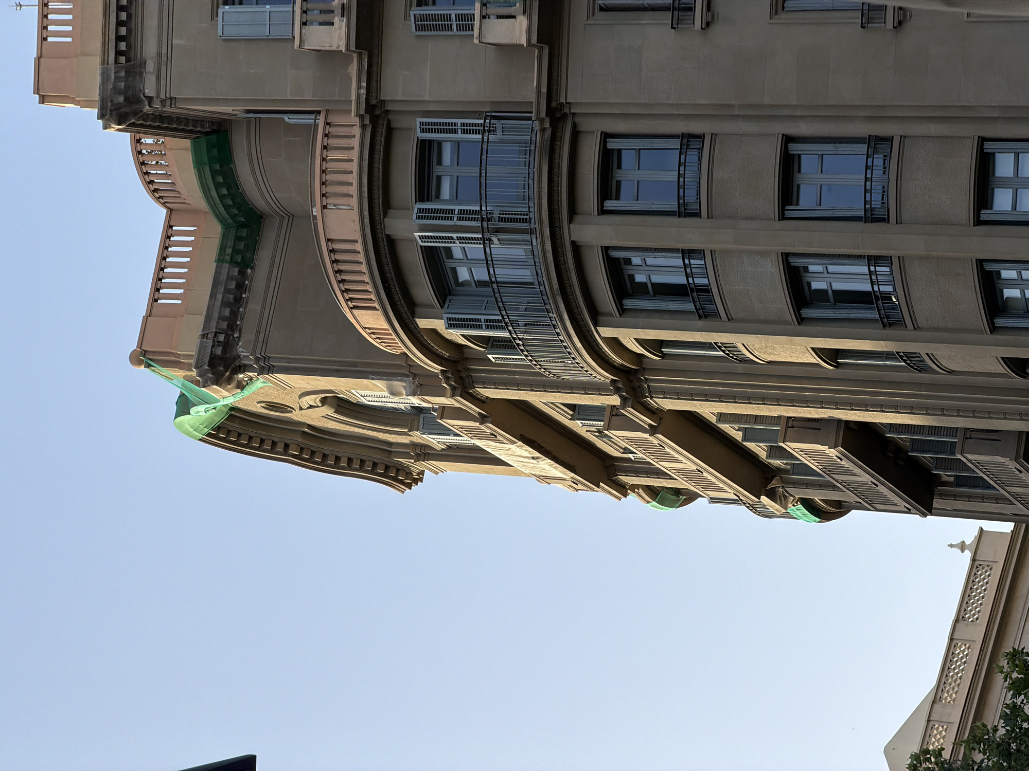 The photograph captures an upward-looking perspective of a light brown stone building in Barcelona, Spain. The building features ornate architectural details, including balconies with railings and several windows with grey shutters. Some sections of the building are partially covered by green netting, suggesting ongoing construction or renovation work. The sky is a clear, pale blue, indicating daytime. To the left of the main building, a portion of another building with a decorative railing and a pointed finial is visible. The sunlight casts shadows on the building's facade, highlighting its textured surface and architectural features.