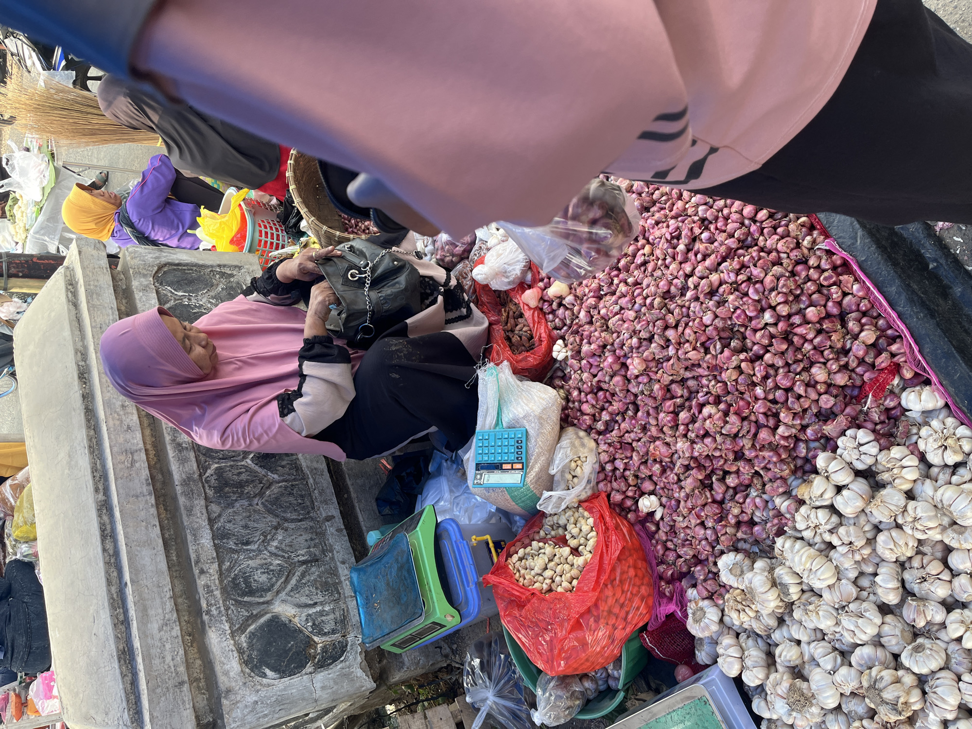 This is a medium shot, eye-level, outdoor photograph taken at a market in Kemuning, Indonesia. The central subject is a woman wearing a pink hijab and a black and pink outfit, sitting behind a stall filled with produce. In front of her, she is sorting through items in a black handbag. To her right, a large pile of red onions spills out onto the ground. To her left, a pile of white garlic bulbs and a red plastic bag filled with what appear to be nuts are visible. A green weighing scale and a light blue calculator are also on the left side of the stall. In the background, other market-goers can be seen, including a woman in a purple hijab and another in a dark hijab, and various goods are displayed. The ground surface is rough and uneven, suggesting a typical outdoor market environment. The lighting indicates it is daytime, possibly late morning or afternoon, with harsh shadows.