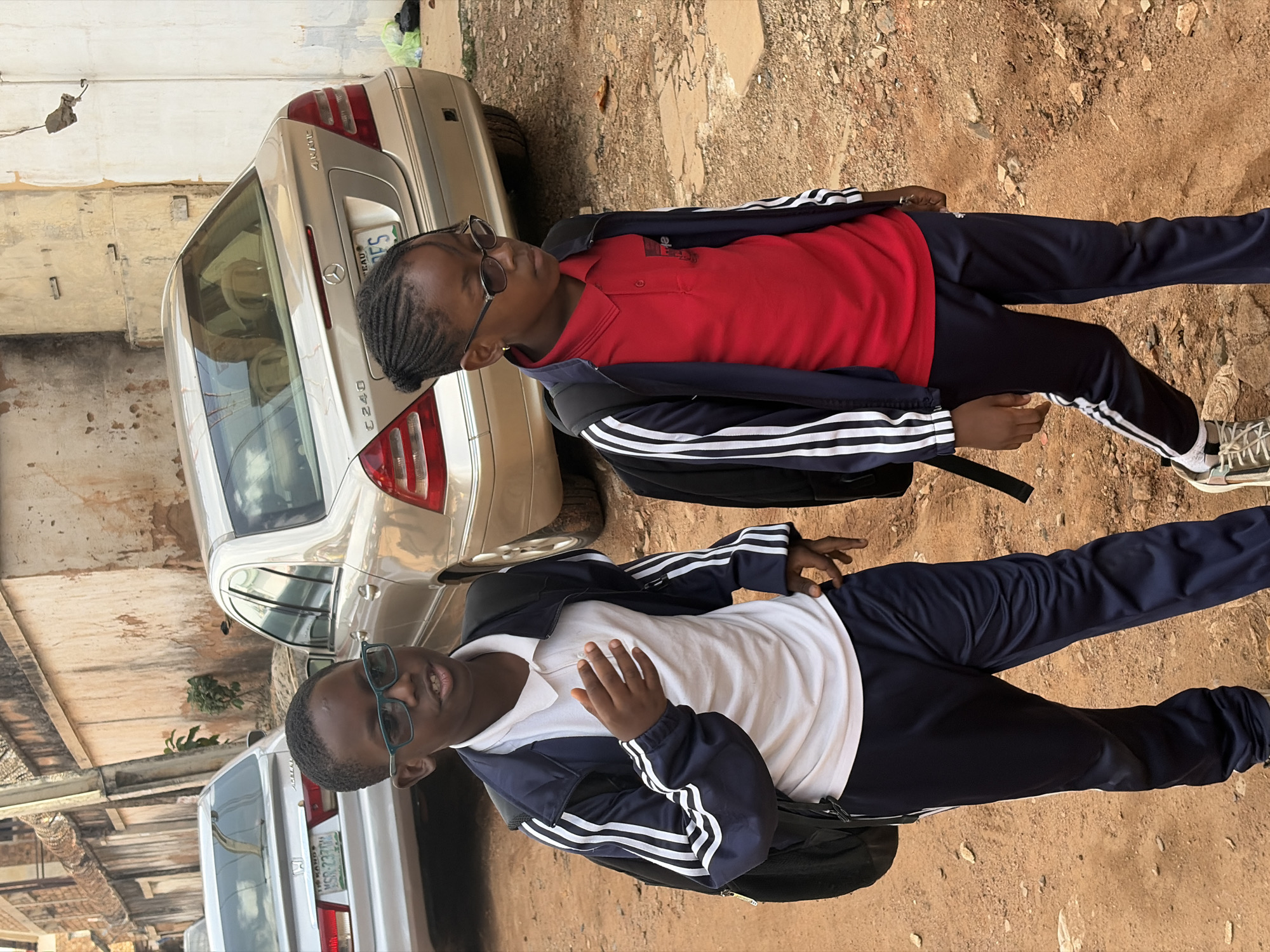 Two young Black individuals, likely children, are standing outdoors next to a silver Mercedes-Benz C240 sedan. The individuals are dressed in athletic tracksuits, one in a red polo shirt and navy blue track pants with white stripes, and the other in a white polo shirt with a navy blue track jacket and navy blue track pants with white stripes. Both are wearing sneakers and sunglasses.
The setting appears to be a street or a yard with a dirt and gravel ground. In the background, a wall and other parked vehicles are visible, suggesting an urban or semi-urban environment. The lighting suggests it is daytime, likely sunny given the shadows. The overall mood seems casual and relaxed. The Mercedes-Benz has a Nigerian license plate that is partially obscured. The image is oriented vertically, with the subjects positioned along the right side.