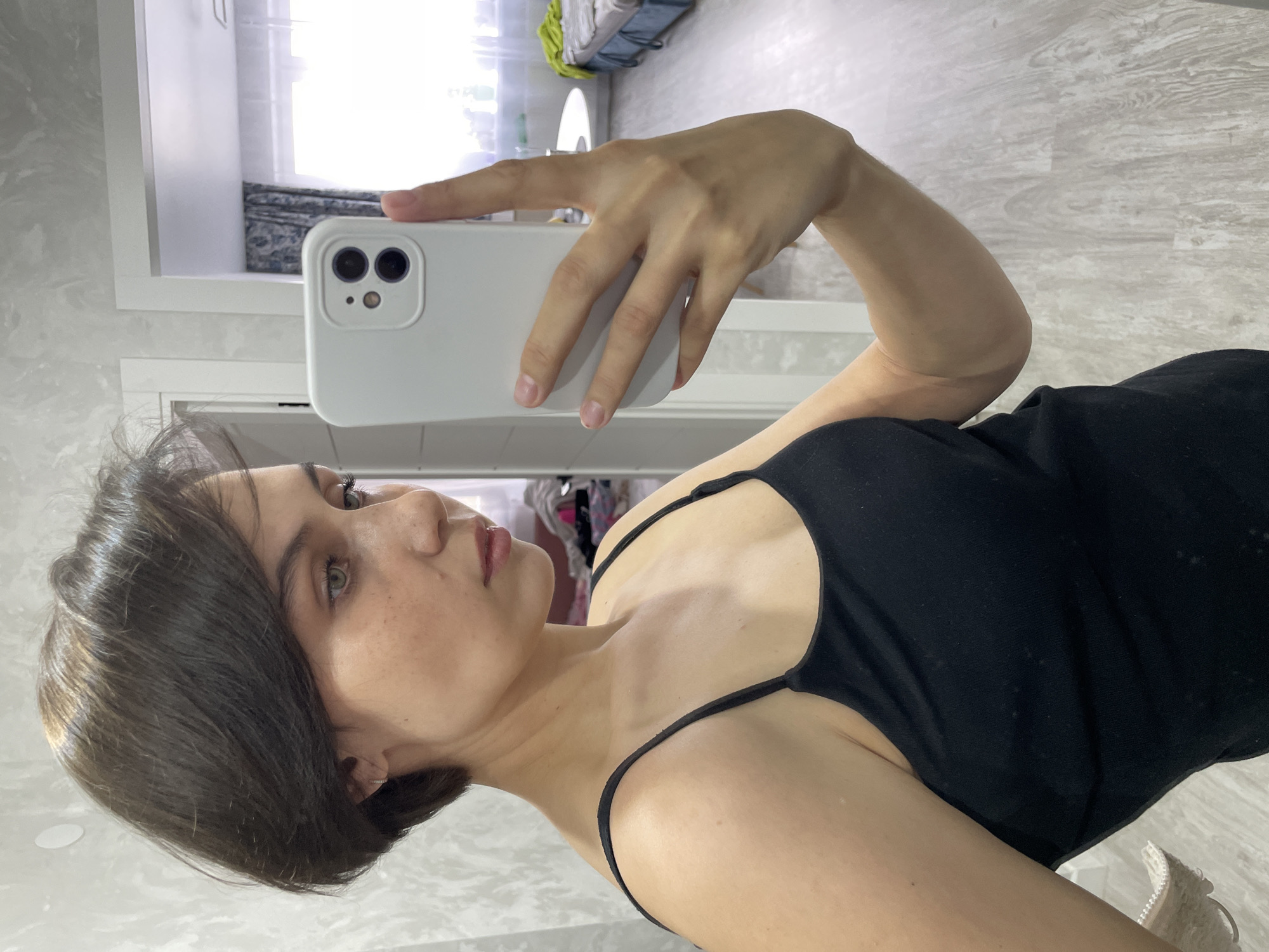 A young woman with short, dark brown hair and light-colored eyes takes a mirror selfie indoors. She is positioned slightly to the right of the frame, looking towards her reflection. Her expression is neutral and calm. She wears a black spaghetti-strap tank top. Her left arm is visible, holding a white smartphone with a light-colored case, which has two main camera lenses and a smaller sensor. Her hand and natural fingernails are visible gripping the phone.
The background reveals a home interior, likely a bedroom or living space, in Izhevsk, Russia. The walls have a light, subtle pattern, and a white door frame is visible behind her. To the upper left, a window lets in bright daylight, with a dark patterned curtain visible through it. The reflection in the mirror also shows what appears to be part of a bed or furniture and some clothes or textiles in the background. The floor has a light-colored wood-grain pattern. Partially visible at the bottom right are white sneakers. The photo is taken during the daytime, indicated by the natural light. There is no visible text.