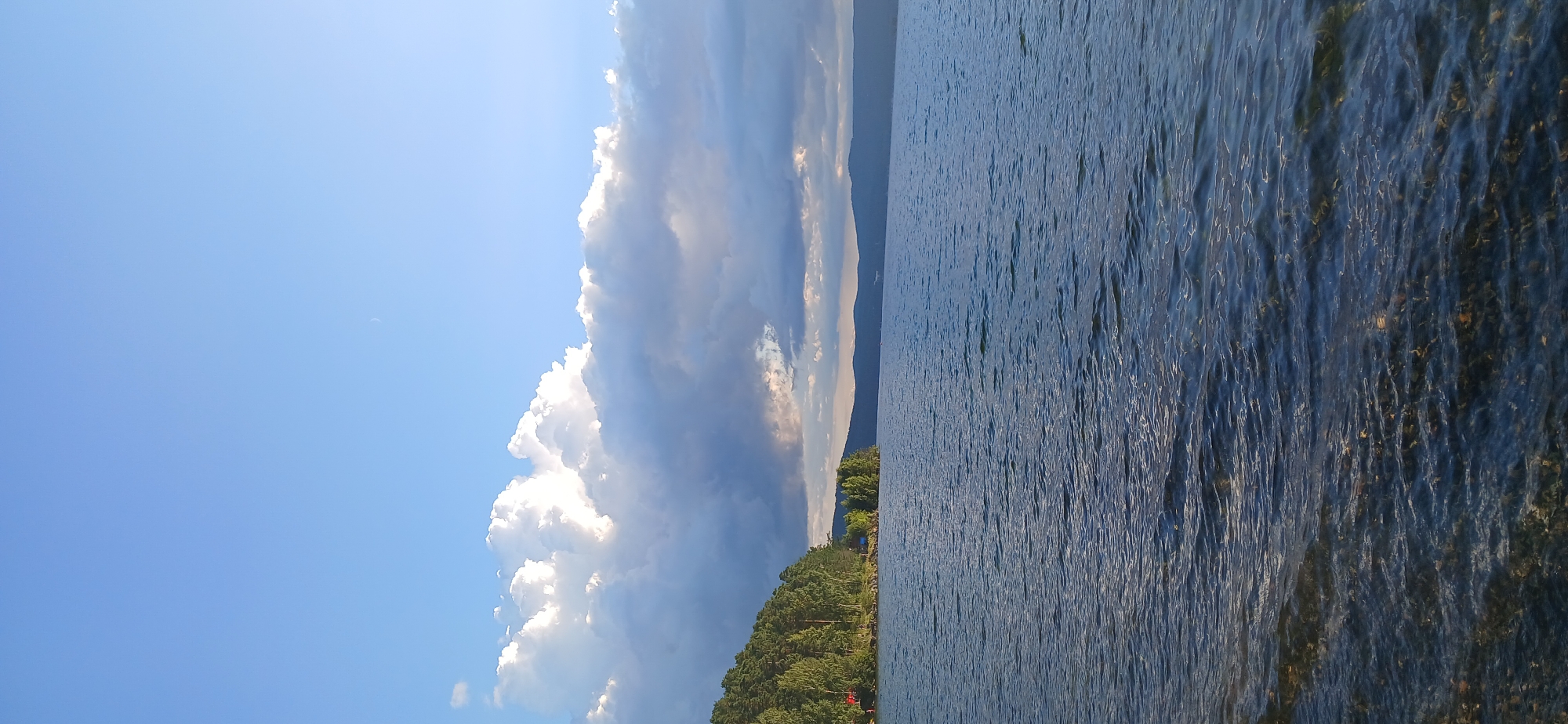 This is a landscape photograph of a lake, likely taken from the shore. The water in the foreground is clear and shows submerged rocks and pebbles. The lake stretches out towards a distant treeline and hills. On the left side of the image, a densely wooded hillside slopes down to the water's edge. Some structures, possibly buildings or tents, are partially visible amongst the trees. The sky above is a bright blue, with a large formation of white, puffy clouds dominating the upper left quadrant. A faint crescent moon is visible in the upper right portion of the sky. The scene suggests a natural, outdoor environment, potentially a recreational area at Lake Turgoyak, Russia, known for its clear water and surrounding forests. There are no visible people in the frame. The overall impression is one of tranquility and natural beauty.