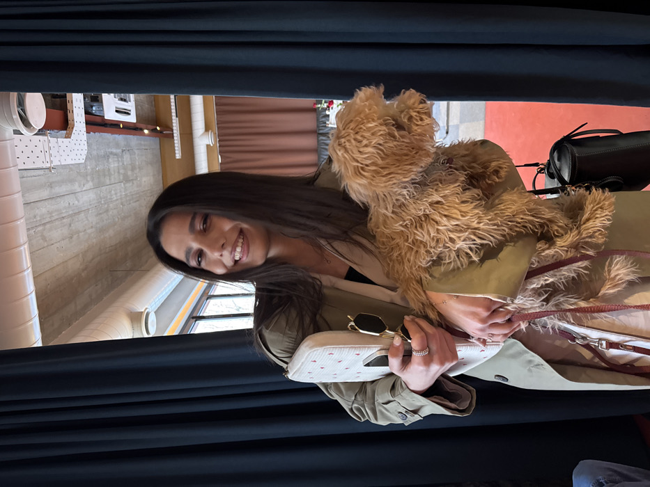 The image depicts a young adult female inside Sherep Restaurant, located at Amiryan St. 1, Yerevan 0001, Armenia. She is positioned centrally, smiling broadly at the camera. The woman wears a light olive-green trench coat over light-colored pants and a dark inner garment. Her long dark hair is down. In her left arm, she cradles a white clutch bag adorned with small red patterns, into which a pair of gold-framed sunglasses is tucked. Her left hand also holds a dark red dog leash. In her right arm, she holds a small, light-brown, curly-haired dog that faces away from the camera. A dark-colored cylindrical crossbody bag hangs from her right shoulder. The background reveals an interior space with a concrete ceiling featuring exposed ventilation ducts and recessed lighting. Dark blue curtains frame both sides of the subject, with lighter brown curtains and windows visible further in the background to the right and left, respectively. A section of red flooring or carpet is visible in the lower right.