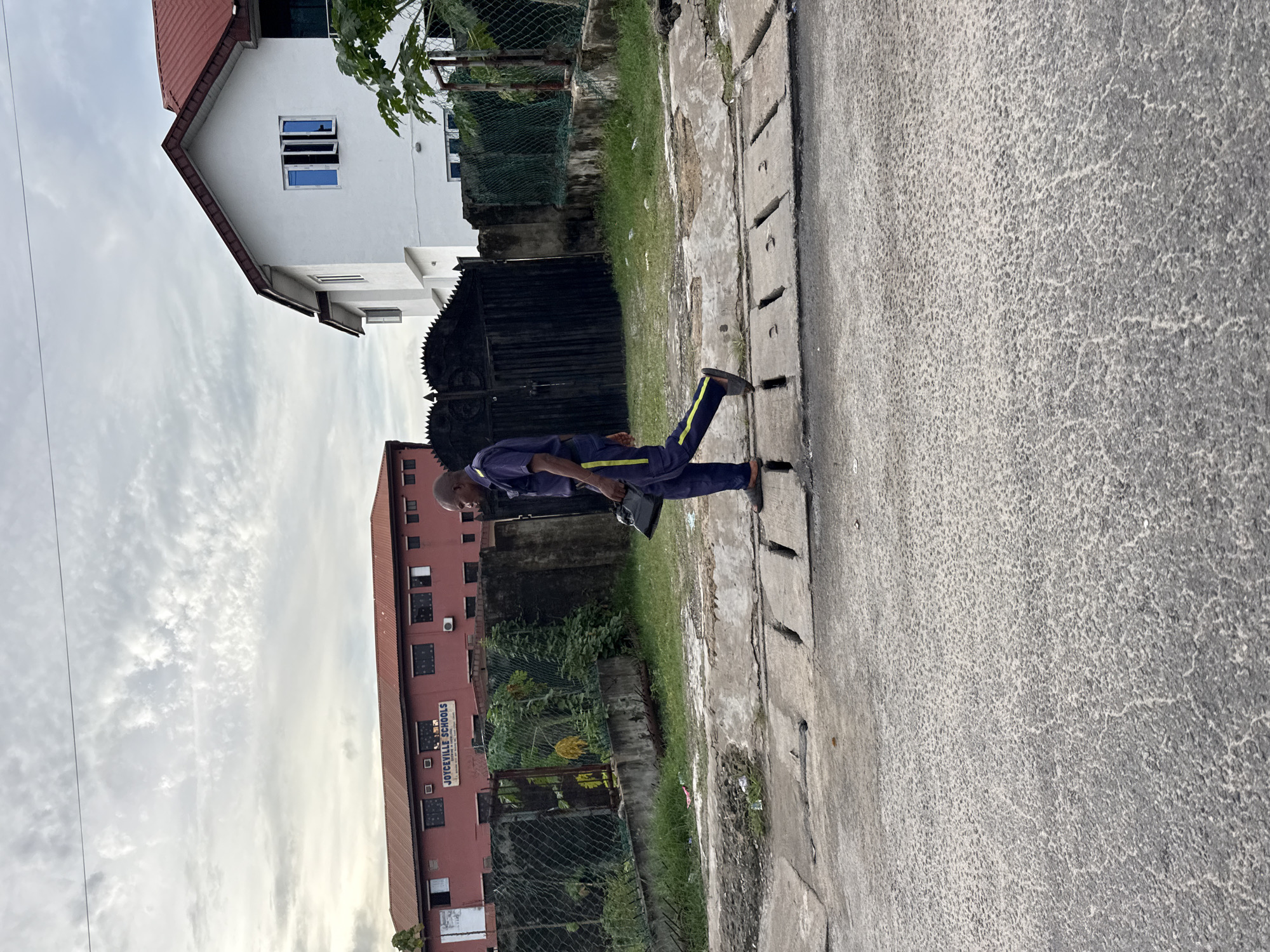 A middle-aged, balding man wearing a navy blue tracksuit with yellow accents walks along a concrete sidewalk adjacent to a gray asphalt road in Ado, Nigeria. He carries a black rectangular object in his left hand. To his left is a green wire fence, behind which are buildings. One building, with a red facade, has a sign reading "JOYCEVILLE SCHOOLS". A white house with a red roof is also visible. The sky is cloudy.