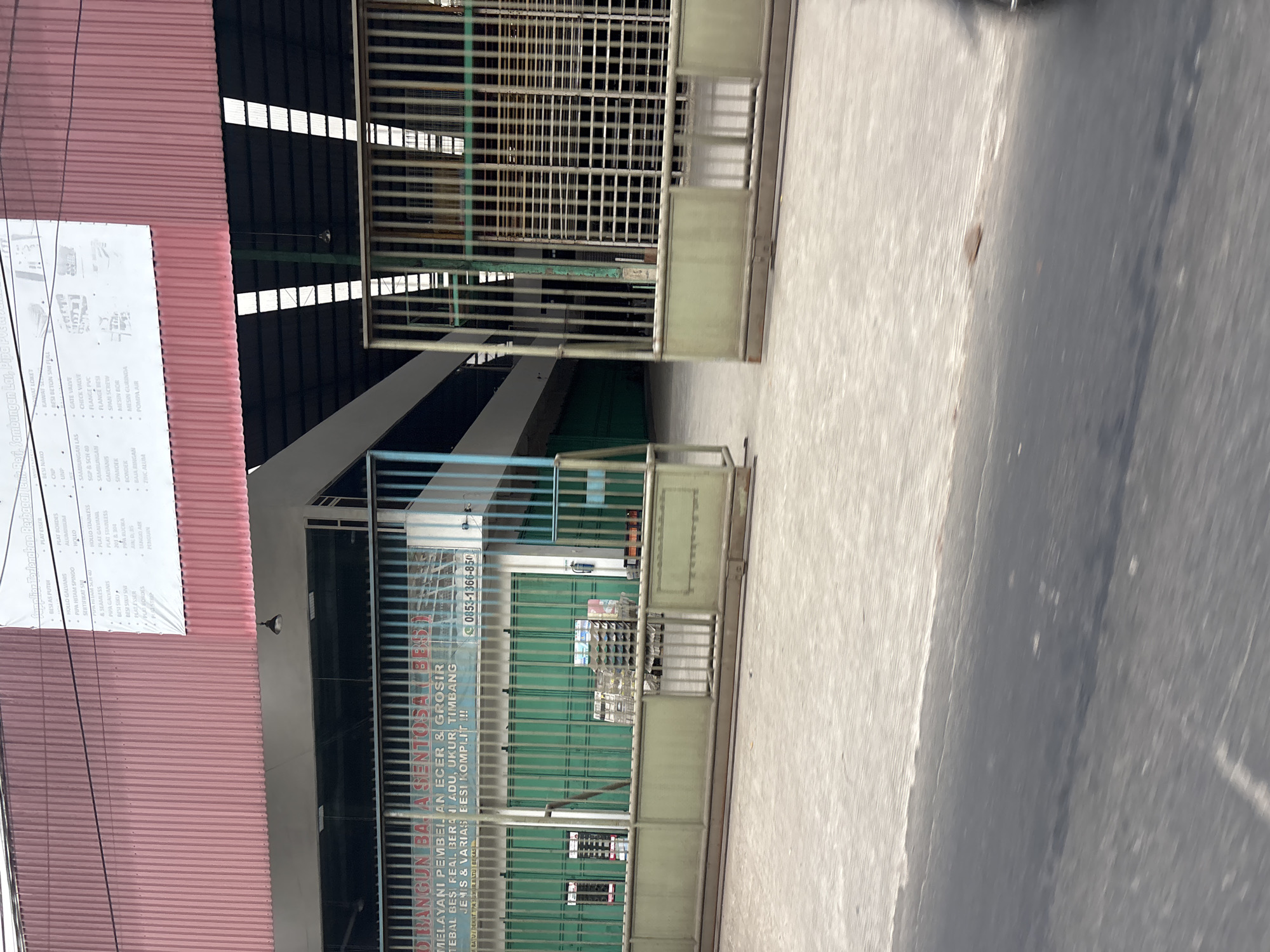 The image shows the exterior of a building in Bekasi, Indonesia. A sign above a large metal gate reads "BANGUN BAJA SENTOSA (BBS)" and indicates that they provide retail and wholesale services for iron, steel, and other materials, with a phone number of 0853-1366-850. The building has a red corrugated metal roof and large windows. The gate is open, revealing a dimly lit interior with stacks of materials and a conveyor belt. The foreground shows a paved area and a street with some motion blur, suggesting the photo was taken from a moving vehicle. The overall scene depicts a commercial establishment, likely a hardware or construction materials supplier, in an industrial or commercial district of Bekasi.