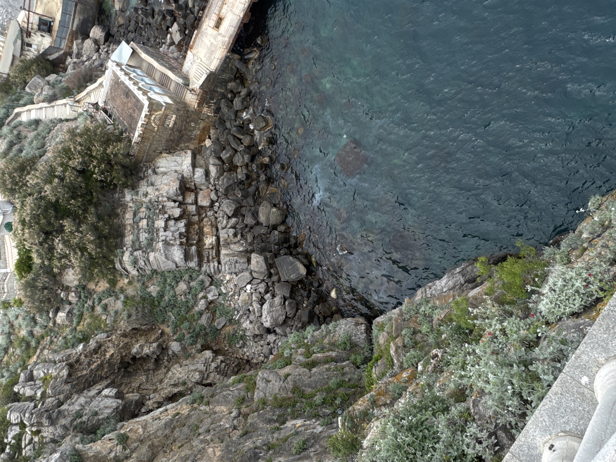 A high-angle shot depicts a rocky cliffside meeting the water's edge in Гаспринский поселковый совет, Ukraine. Buildings are perched precariously on the cliff, with stone structures and walkways visible. The water is a deep blue-green, with rocky formations underwater and along the shoreline. Vegetation clings to the rocks, consisting of shrubs and greenery. The photograph has a slightly rotated perspective, emphasizing the steepness of the terrain.