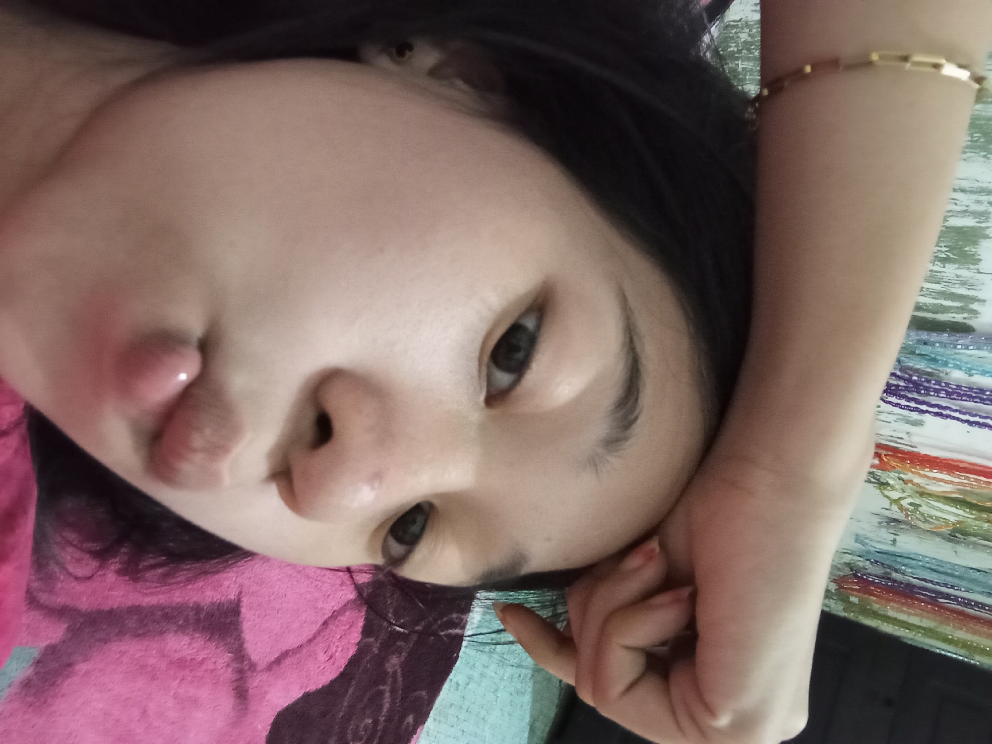 The image shows a close-up of a young woman's face, with her arm resting on her forehead. She is wearing a gold bracelet on her wrist. Her lips are pursed, as if blowing a kiss. The background appears to be a bedroom, with a pink blanket and a wall decorated with beads. The image is taken from a selfie perspective. No specific location in Rejosari, Indonesia is visible or inferable from the image.