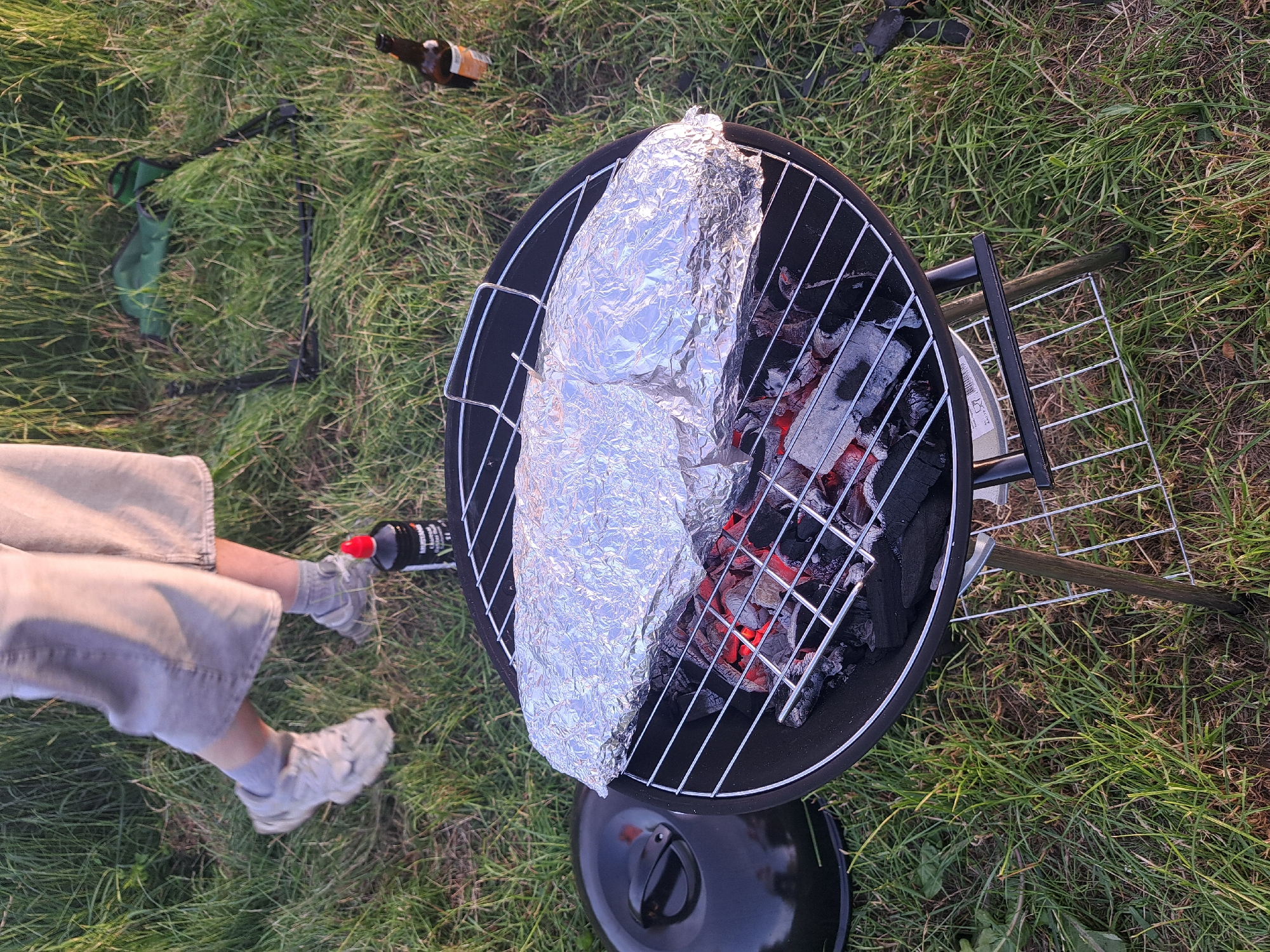 In a grassy, outdoor setting, a person is preparing food on a barbecue grill. The grill is a black, circular charcoal barbecue with a metal grate. On the grate are two large items wrapped in aluminum foil, presumably food being cooked. Below the grate, glowing red coals are visible, indicating the grill is hot.
To the left of the grill, a person's legs and feet are visible. They are wearing light-colored shorts, grey socks, and light-colored athletic shoes. The person's right hand is holding a small, black bottle with a red cap, possibly a sauce or seasoning. In the background, a green fabric object is on the grass, and a brown glass bottle lies nearby.
The scene appears to be taking place outdoors, likely in a park or backyard, given the grass and natural light. The lighting suggests it is either early morning or late afternoon, with the sun casting long shadows and illuminating the scene with a warm glow. The weather seems pleasant, as the person is dressed in shorts and the setting is outdoors. The overall atmosphere suggests a casual, leisurely activity like a picnic or camping trip. No specific Latvian landmarks are visible, but the location context indicates it is Riga, Latvia.