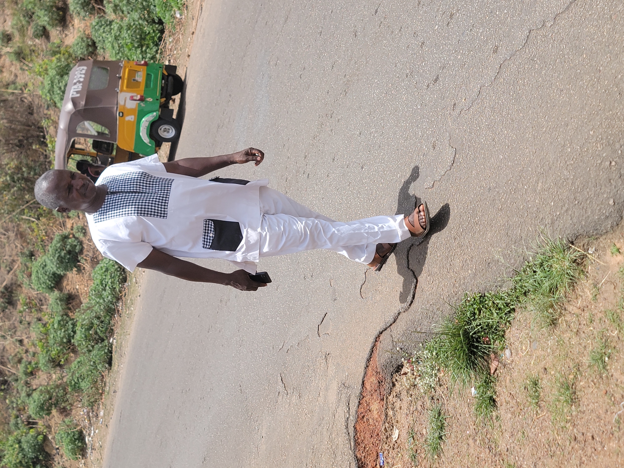 A man wearing a white, long-sleeved tunic-style shirt with a black and white checkered pattern on the chest, white pants, and brown sandals is walking on a cracked asphalt road. He holds a black object in his left hand. A three-wheeled vehicle is parked on the shoulder of the road. Vegetation and a hill are on the left side of the road. The scene is located on a street in Jos, Nigeria.