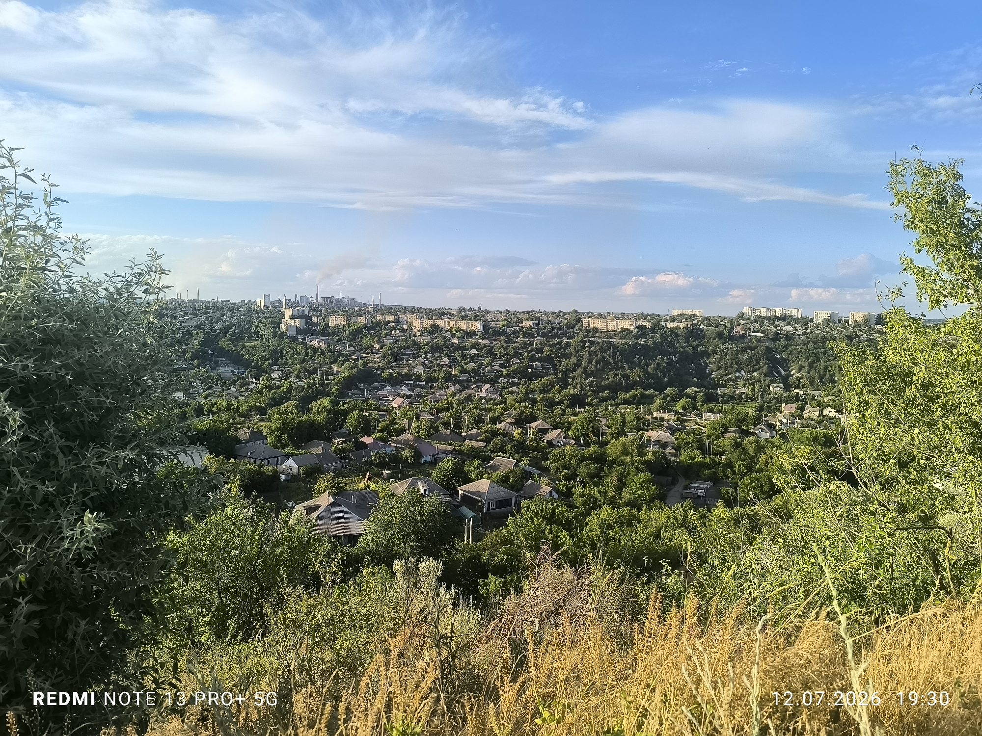 The image shows an expansive view of a city, likely taken from a high vantage point overlooking a valley. The foreground is dominated by dry, golden grasses and the lush green foliage of trees and bushes. In the middle ground, a dense residential area with numerous houses and rooftops is nestled within a verdant landscape. Beyond the residential area, the city extends further, with visible industrial structures, including tall chimneys emitting smoke, and a cluster of taller apartment buildings. The sky above is a bright blue, dotted with wispy white clouds, suggesting fair weather. The overall impression is one of a sprawling urban settlement surrounded by nature. The bottom left corner displays "REDMI NOTE 13 PRO+ 5G" and the bottom right corner shows the date and time "12.07.2026 19:30".