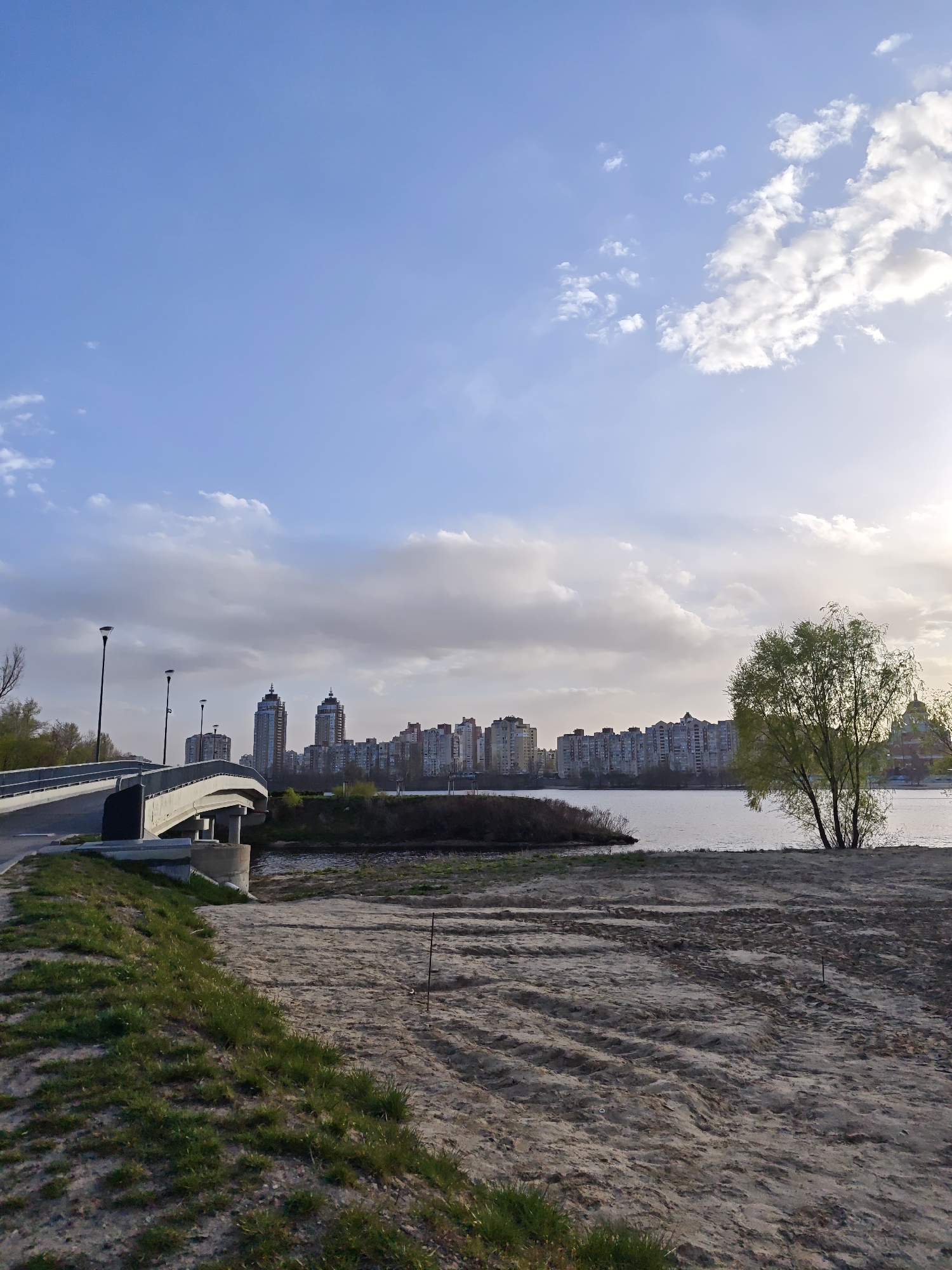 The image presents a serene waterfront cityscape in Kyiv, Ukraine, under a partly cloudy sky. The scene is illuminated by the soft, diffused light of what appears to be late afternoon or early morning, with the sun casting a gentle glow on the clouds and the distant buildings.
In the foreground, a sloping bank composed of light-colored sand or dirt dominates the lower right, with distinct tire tracks or ground patterns visible across its surface. Patches of bright green grass grow on the left side of the bank, indicating lush vegetation.
The midground features a modern, curving concrete bridge or overpass on the left, supported by sturdy piers. It has dark railings and multiple streetlights along its span. This bridge crosses a narrow inlet that connects to a larger body of water, likely the Dnipro River. On the right, a solitary deciduous tree with vibrant green foliage stands by the water's edge, its leaves indicating spring or early summer.
Across the wide expanse of water, a prominent city skyline unfolds. Numerous residential or commercial buildings, varying in height and architectural style, line the distant shore. Two taller, more distinct high-rises are visible towards the center-left of the skyline. The sky above is a blend of clear blue patches and scattered white and grey cumulus clouds, some brightly lit by the low-lying sun on the right.
No people are visible in the tranquil scene, and there is no discernible text. The image captures a quiet moment in an urban natural landscape.