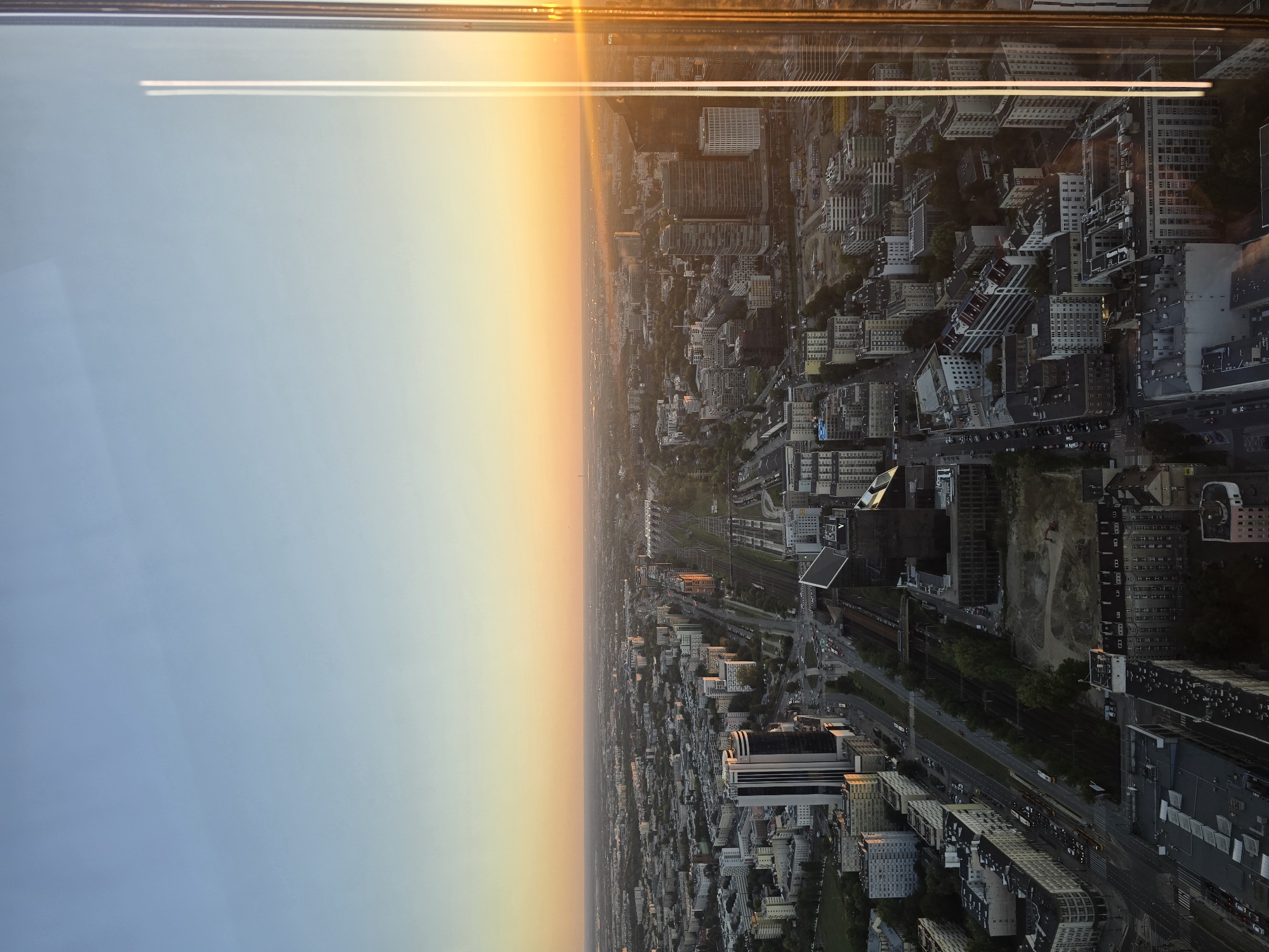 This is an aerial, high-angle photograph of the city of Warsaw, Poland, taken at sunset. The sky is a gradient of pale blue to warm orange, indicating the sun is low on the horizon. A bright, golden sun flare is visible on the right side of the image, reflecting off the glass of the viewpoint.
The cityscape below is dense with buildings of varying heights and architectural styles, suggesting a mix of modern and older construction. Prominent features include a large, modern building with a dark facade in the mid-ground, and a long stretch of railway tracks cutting through the urban environment. The tracks are flanked by numerous buildings, roads with traffic, and areas of green space. In the foreground, more residential or office buildings are visible. The overall impression is of a bustling, developed urban area at the end of the day. The specific location within Warsaw is difficult to determine with certainty without further information, but the density of high-rise buildings and the presence of significant railway infrastructure suggest a central business district or a major transportation hub.