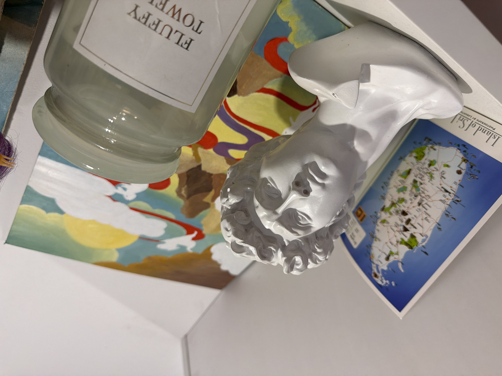 The image depicts a close-up, high-angle shot of objects arranged on what appears to be a shelf or tabletop. The items include a glass jar with a label reading "FLUFFY TOWEL" in capital letters, a white plaster bust of a person, and a postcard or book cover displaying a map of an island with the text "Island of..." partially visible. The setting appears to be indoors, likely in Большая Шильна, Russia, but specific location details are not discernible from the image.