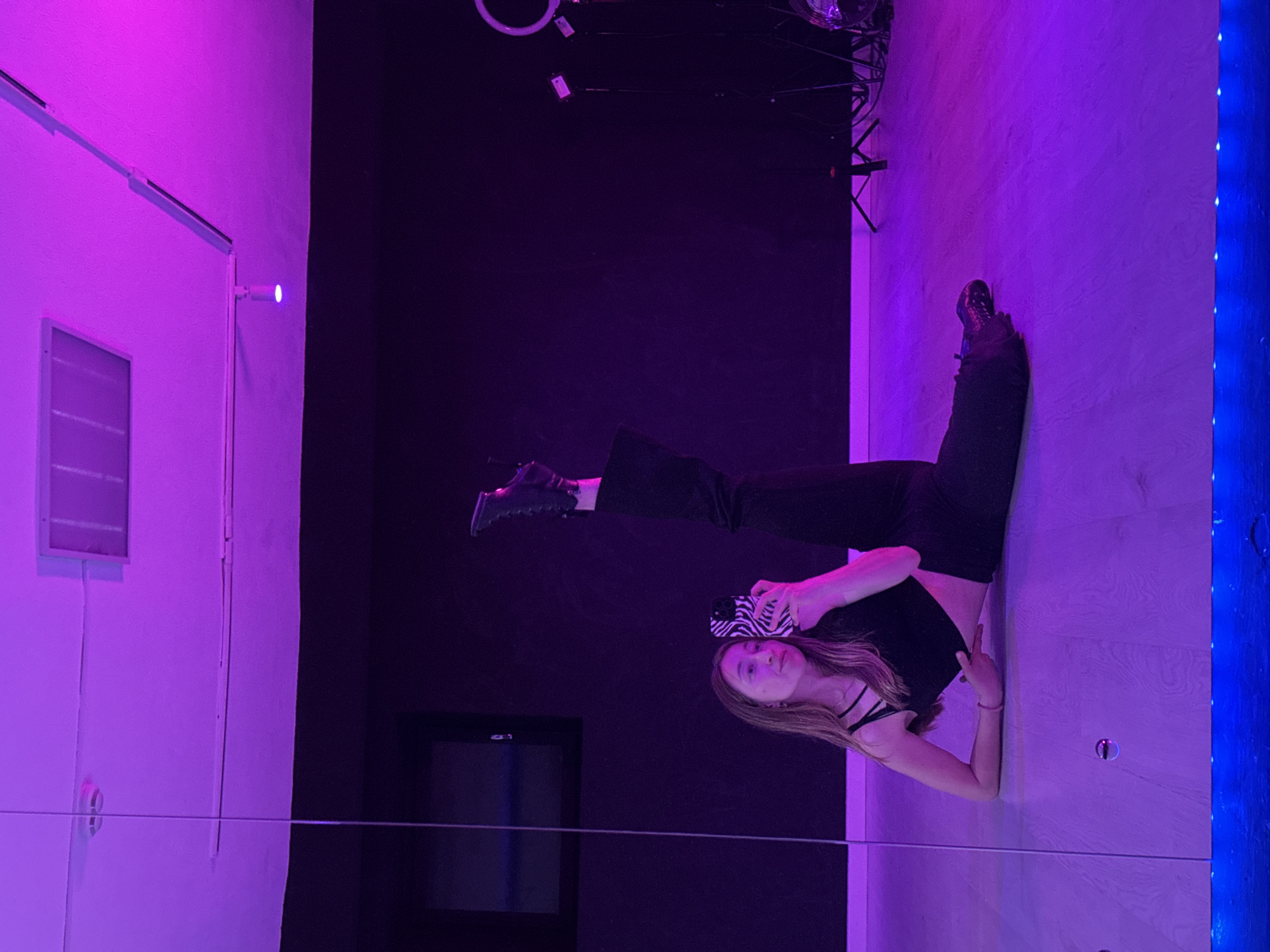A young woman in black pants and a black top is lying on her side in a brightly lit room, possibly in Chaikovsky, Russia. She is holding a zebra-print phone in her right hand and appears to be taking a picture or video of herself. Her head is tilted slightly towards the camera, and her left arm is extended to the side. The room has a distinct purple and blue lighting scheme. To the left is a white wall with a window, and to the right is a wall with blue LED lights. In the background, there's a dark wall with some lighting fixtures and equipment visible near the top. The floor has a wood-like texture. There's a reflective surface, presumably a mirror, at the bottom of the frame, reflecting the scene.