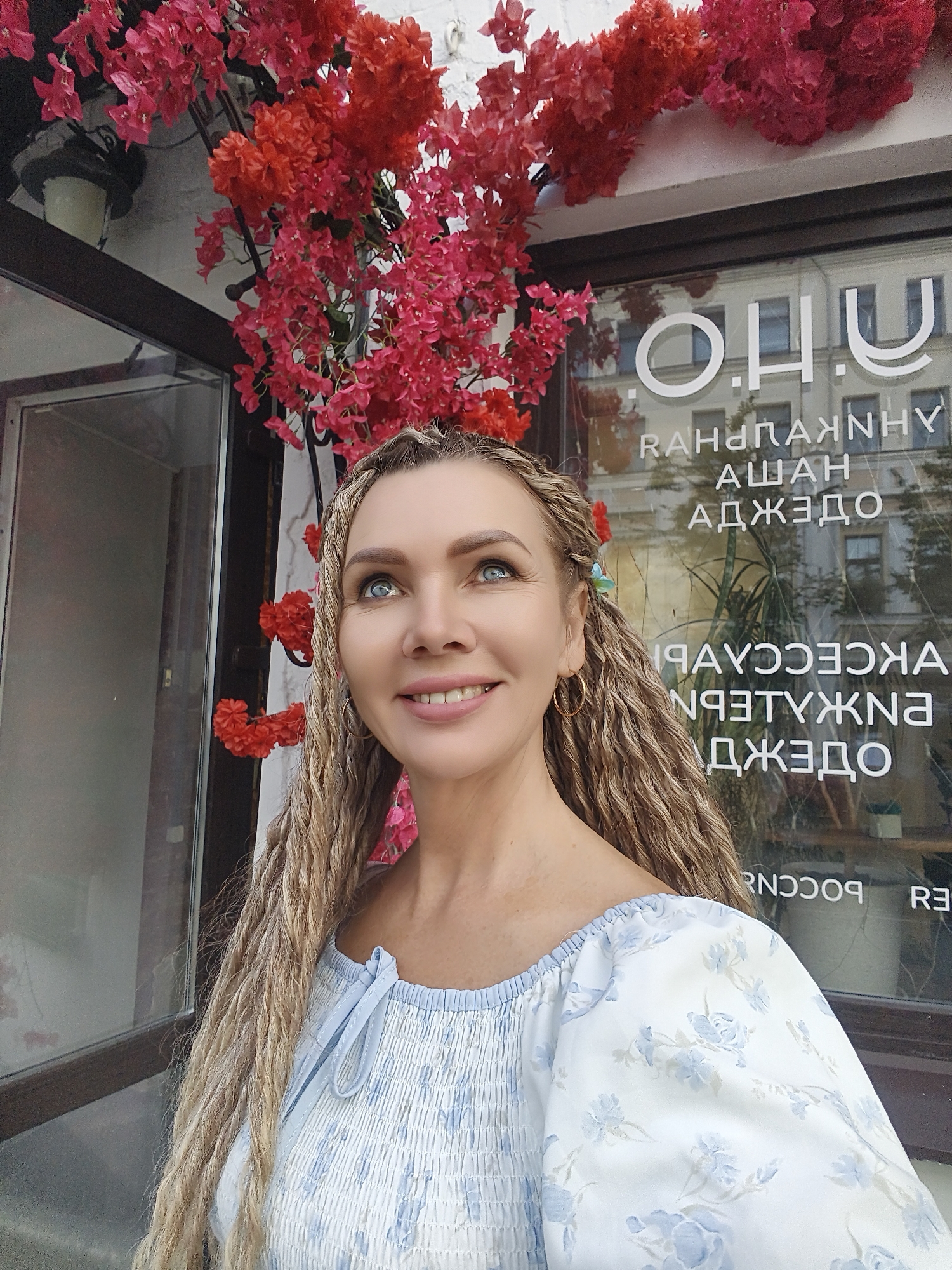 A woman with blonde dreadlocks and blue eyes looks up and smiles in front of a storefront. She is wearing a blue and white floral dress. The storefront has large windows with text visible, some of which is reversed due to reflection. Red and pink flowers hang above the storefront.