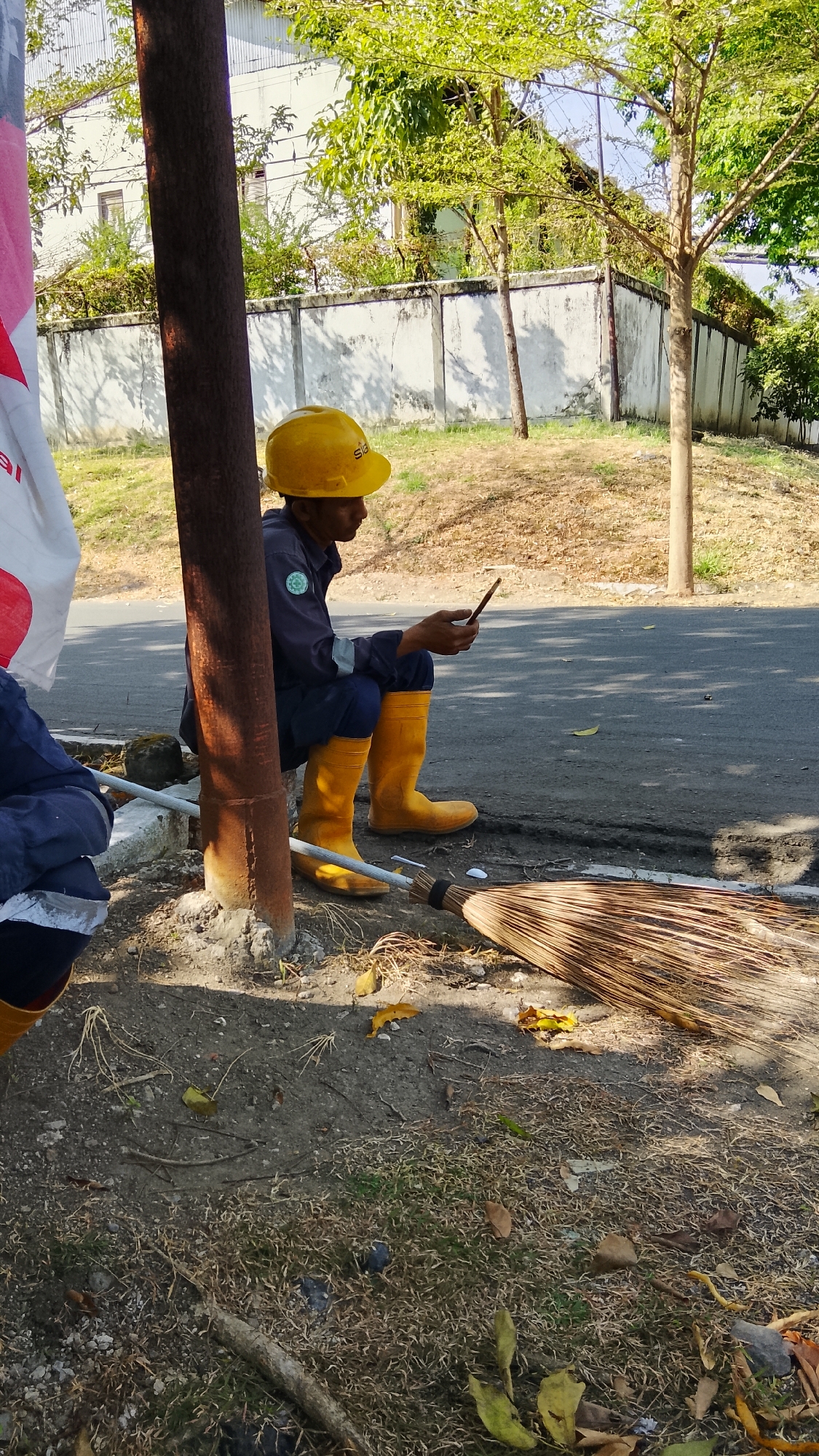 A person wearing a yellow hard hat, dark blue uniform, and yellow rubber boots is sitting on the ground next to a metal pole, looking at their mobile phone. A broom made of natural fibers lies on the ground in front of them. The scene appears to be outdoors, possibly in an industrial or work area, with a concrete wall and trees in the background. The ground is covered with dirt, dry grass, and fallen leaves. A portion of a white and red flag or banner is visible on the left side of the image. The location is specified as Tlogobendung, Indonesia.