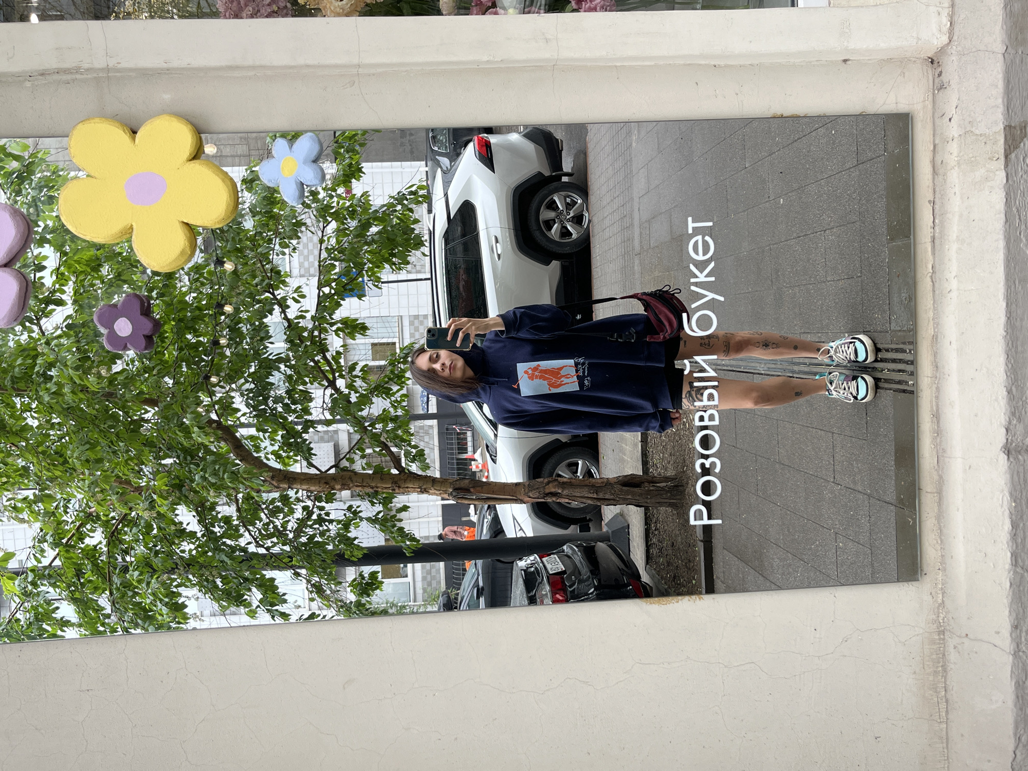 A young woman with dark hair pulled back is taking a selfie in a large, rectangular mirror mounted outdoors. She is wearing a navy blue hooded sweatshirt with an orange graphic of a horse, black shorts, and multi-colored sneakers. A small maroon bag is slung across her body. The mirror reflects a street scene, including a tree with lush green leaves, several parked cars, and a building in the background. The ground is paved with grey tiles. The words "Розовый букет" (Pink bouquet) are visible in white lettering on the mirror, indicating the establishment's name. The overall impression is a casual street-style photograph. The location is Krasnoyarsk, Russia.