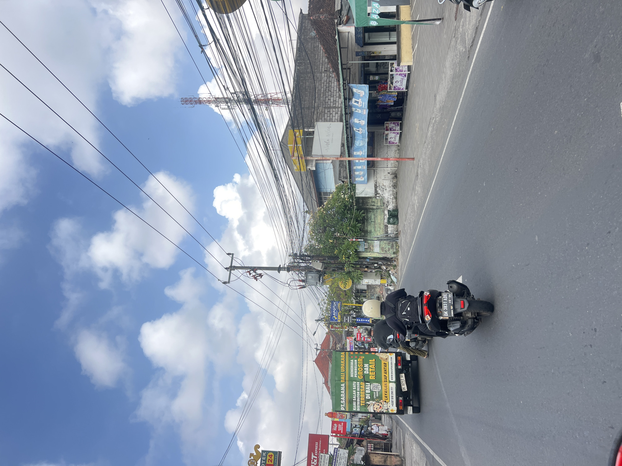 This image captures a street scene in Denpasar, Indonesia, on a cloudy day with a bright sky. The perspective is from a moving vehicle, looking forward. In the foreground, an asphalt road with a white dividing line leads into the distance. Two motorcyclists are visible, one ahead and slightly to the right, wearing a white helmet and black clothing, and another further ahead and to the left, also on a motorcycle.
A white delivery truck with green lettering is in the middle distance, displaying "PT. SARANA BALI UPAKAR", and an Indonesian license plate "DK 8793 AR". Signs for "APOTEK 24 JAM" (24-hour pharmacy) and "T&T Express" are visible on the left side of the road, along with other businesses and buildings.
On the right side of the road, a row of shops features awnings and displays of clothing. A tall telecommunications tower stands prominently in the background. The street is lined with utility poles and a dense network of electrical wires, typical of urban Indonesian infrastructure. The overall atmosphere suggests a busy commercial area.