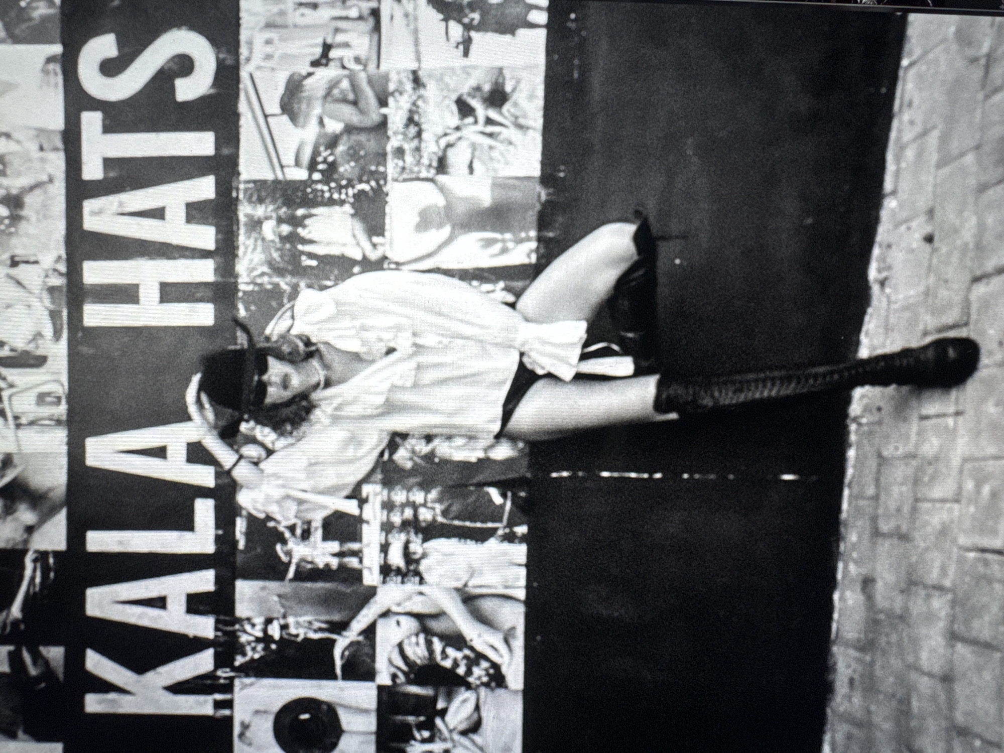 The image is a black and white photograph that appears to be of a magazine or poster. The words "KALA HATS" are vertically oriented on the left. A woman with a long-sleeved white shirt, black shorts, and high black boots is positioned diagonally across the image. Her body is partially obscured by the vertical text on the left side, where there are collage-like images of other people and scenes. The right side of the image consists of a large black vertical section with a brick wall at the edge. There is no specific visible indication of the location being Mengwi, Indonesia.