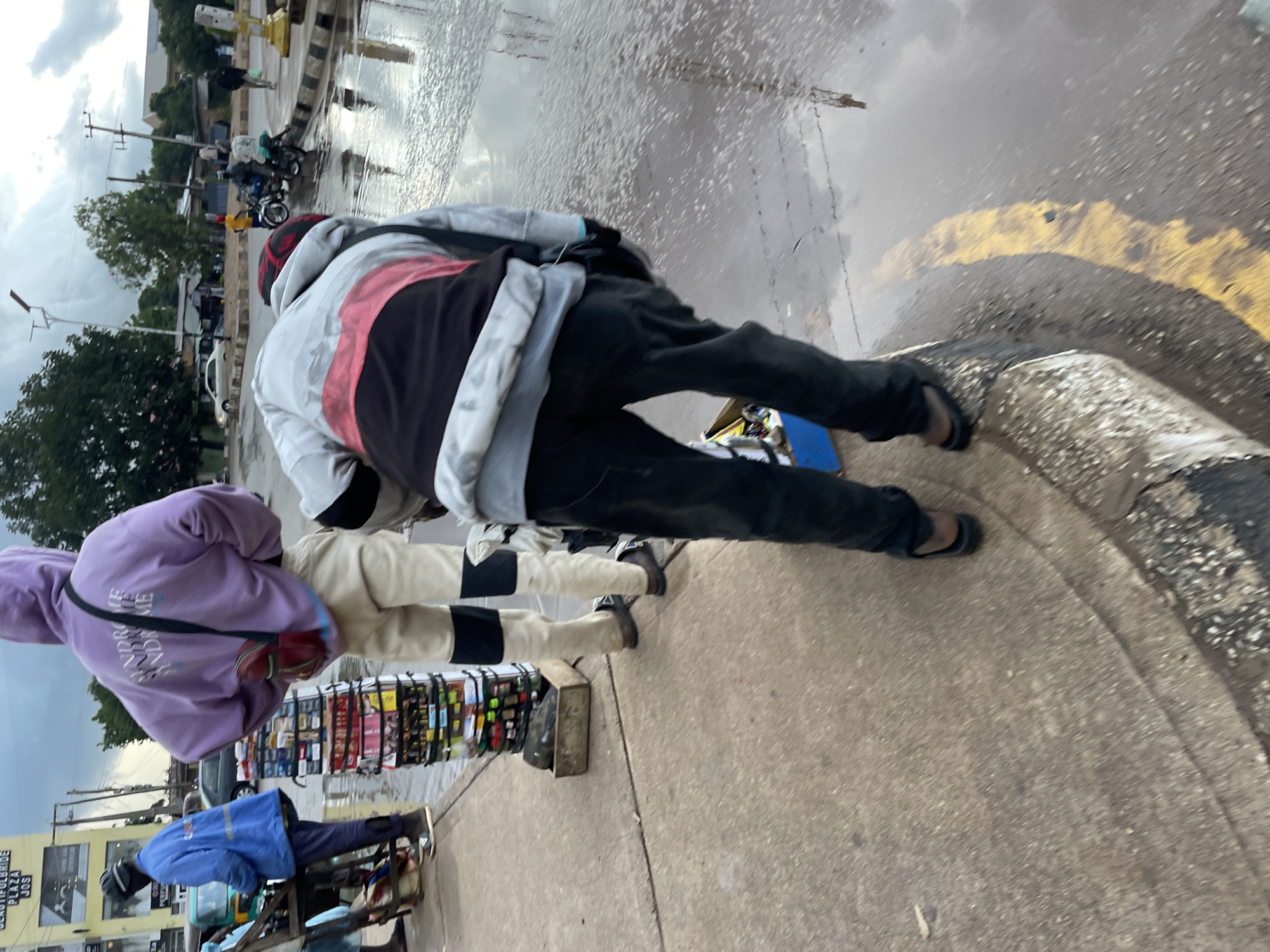 The image depicts a street scene in Jos, Nigeria, following a rainfall. A young person wearing a light purple hoodie with "SYNDROME SYNDROME SYNDROME" printed on the back, paired with light-colored trousers and dark footwear, stands next to a tall stack of items, possibly goods for sale. To the left, another individual in a bright blue jacket and dark trousers is visible, appearing to be tending to a stall or cart with various products. In the background, several motorcycles and a few cars are present on the wet road, reflecting the overcast sky. The ground is wet and shows puddles, indicating recent rain. The overall environment suggests a busy, possibly market-like, urban setting.
