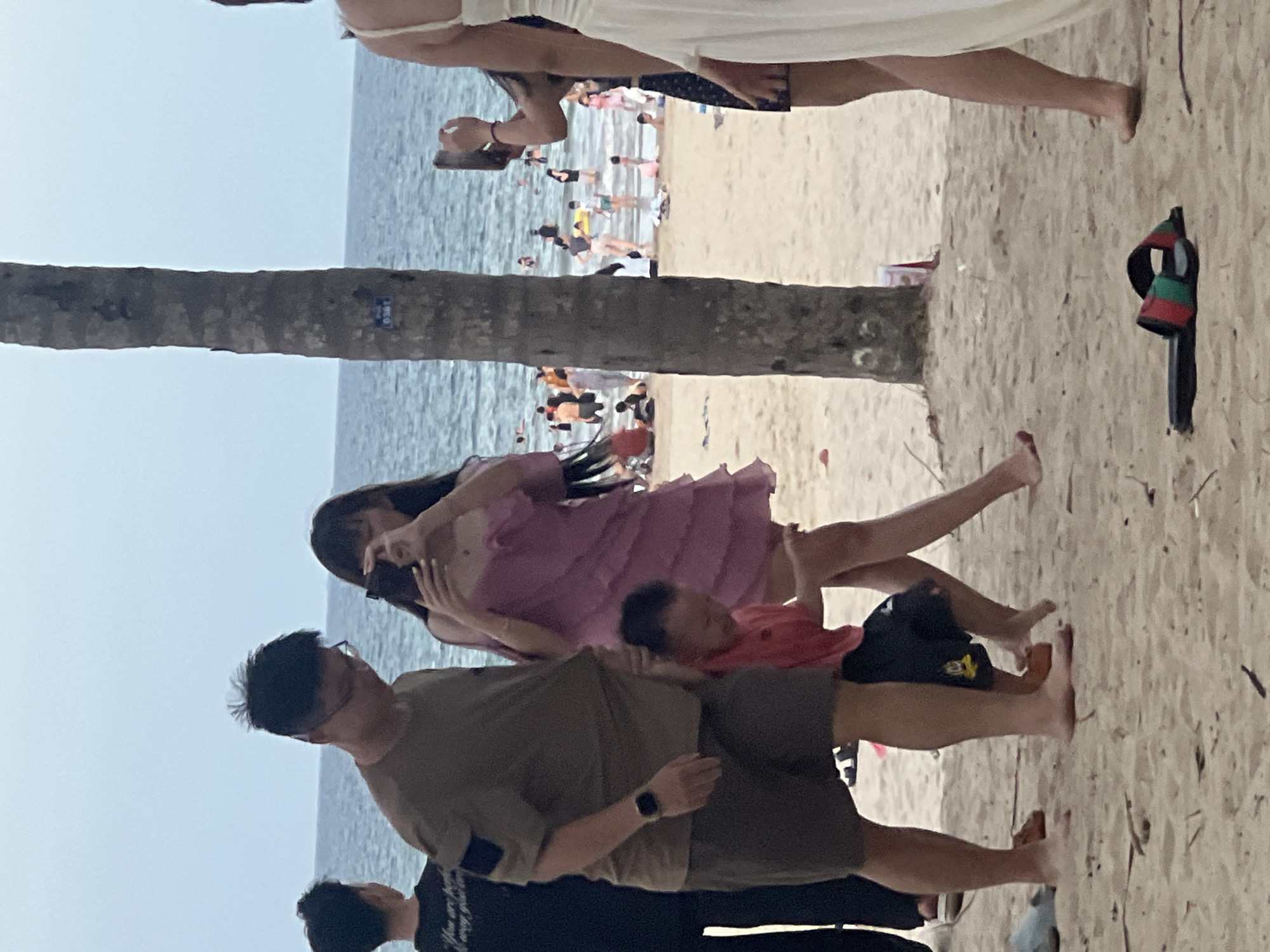 The image depicts a beach scene in Đà Nẵng, Vietnam. The photo is oriented vertically, with the horizon line running horizontally across the image.
In the foreground, three people are visible. A man wearing glasses and a khaki-colored t-shirt is in the center, with a watch on his left wrist. To his left, another person wearing a black shirt is partially visible. To the man's right, a woman in a pink dress is holding a baby in a red shirt. Her hair is dark and long, and she appears to be taking a photo with her phone. The baby is resting on her shoulder.
Behind these individuals, the sandy beach stretches out, with several other people scattered across it. The ocean is visible in the background, with calm waters and a hazy sky, suggesting it might be overcast or early morning/late afternoon. A palm tree trunk is prominently featured on the left side of the image, partially obscuring the view of the beachgoers in the distance. A discarded pair of flip-flops, with red and green stripes, lies on the sand to the right. The overall atmosphere appears relaxed, with people enjoying a day at the beach. There is no visible text in the image.