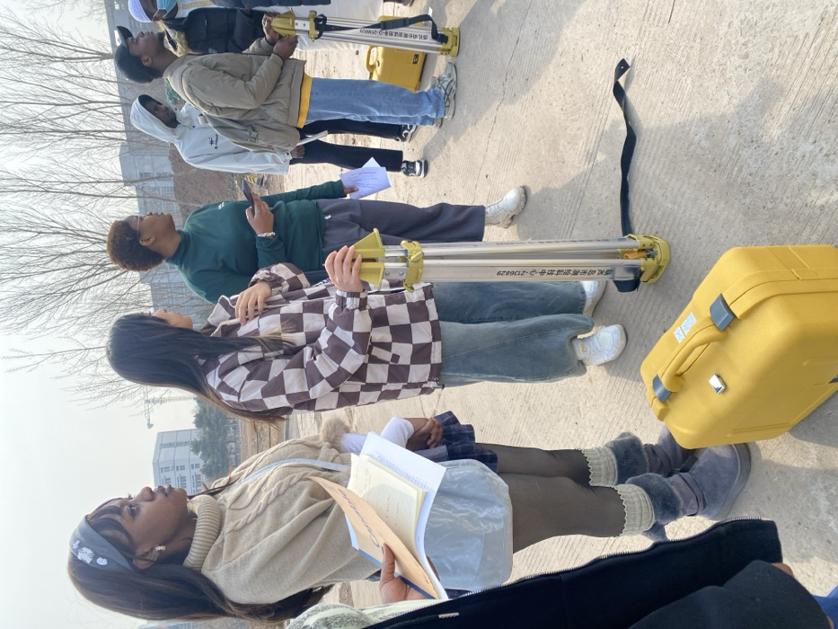 The image captures an outdoor scene in the city of Zhongtun, China, featuring a group of seven individuals standing on a concrete surface under a clear sky. From left to right: a partially visible person in a dark jacket holding papers; a young Black woman with long dark hair, wearing a grey patterned headband, a beige cable-knit turtleneck sweater, a light blue skirt-like garment over dark leggings, and grey boots with knitted cuffs, holding papers and a notebook; a young East Asian woman with long dark hair, wearing a brown and white checkered jacket, blue jeans, and white sneakers, with her right hand resting on a yellow and silver surveying prism pole that stands upright on the ground; a young Black woman with short dark hair, wearing a dark green turtleneck and grey trousers with white boots, holding a smartphone; a young Black man in a light grey hooded sweatshirt, dark pants, and black sneakers, with a backpack; a young Black man wearing a black cap, light brown jacket, yellow shirt, blue jeans, and light grey sneakers, holding a second yellow and silver surveying prism pole; and a partially visible individual in a dark jacket and white face mask. All individuals are oriented towards the right side of the frame. A bright yellow hard case is on the ground in the foreground, and a black strap lies near the first prism pole. The background shows bare trees, several multi-story buildings, and a construction crane, indicating an urban or developing area.