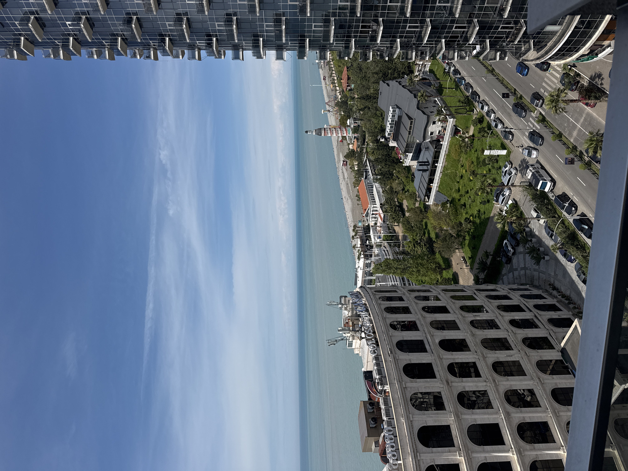 This image depicts a view of Batumi, Georgia, from a high vantage point, possibly a tall building. The scene includes the Black Sea, a beach, and a road running along the coast with vehicles and pedestrians. Several architectural elements are visible, including a building with a distinctive curved facade. The sky is partly cloudy. The image is rotated 90 degrees clockwise.