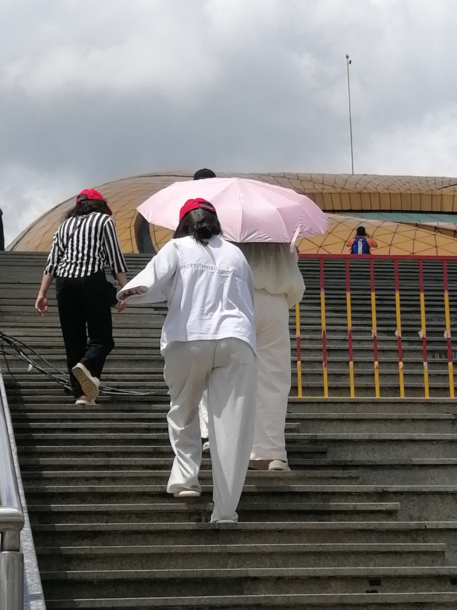 The image depicts a group of individuals ascending a wide, outdoor stone staircase. The sky is overcast with gray clouds. In the foreground, three people are visible from the rear. The person on the left wears a black and white striped long-sleeved shirt, black pants, and a red cap. They are stepping onto a higher stair. To their right, a person in a white long-sleeved shirt and white pants is walking up the stairs, holding a light pink umbrella overhead. Further to the right and slightly behind, another person, also in white clothing and a red cap, walks up the stairs. In the background, a portion of a large, modern building with a golden, curved roof is visible, along with a yellow and red railing. A person wearing a blue backpack is seen on an elevated walkway in the distance. The scene suggests a public outdoor space in Da Lat, Vietnam, possibly a tourist attraction or public building.