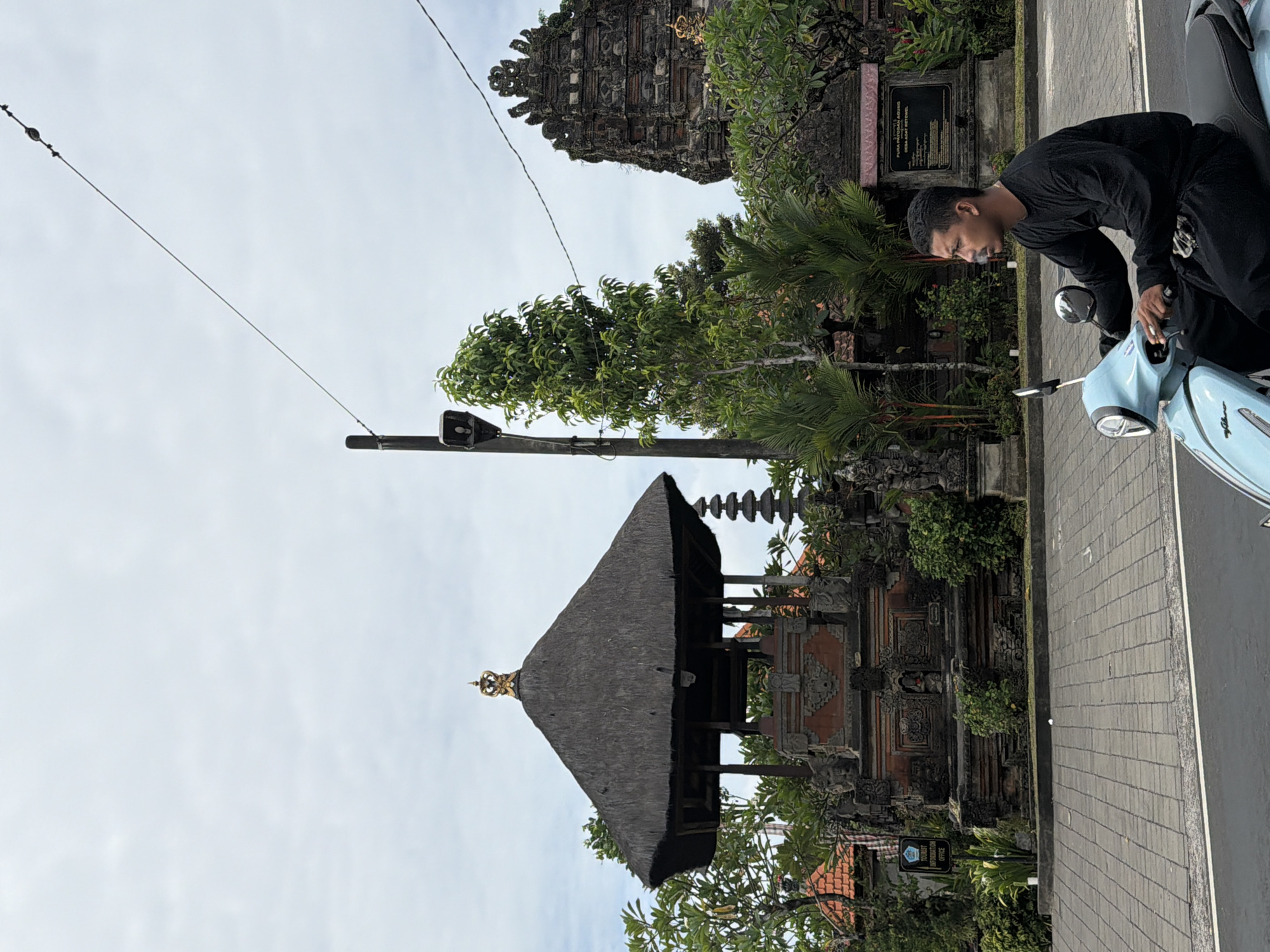 A man wearing a black long-sleeved shirt and black pants rides a light blue scooter with a black seat, smoking. The scooter is on a grey paved road with a white stripe. In the background, there is a traditional Balinese temple with a thatched roof and intricate stone carvings. A tall, dark stone tower with multiple tiers is also visible. Green trees and foliage surround the temple. The sky is overcast. The scene appears to be in Sukawati, Indonesia.
