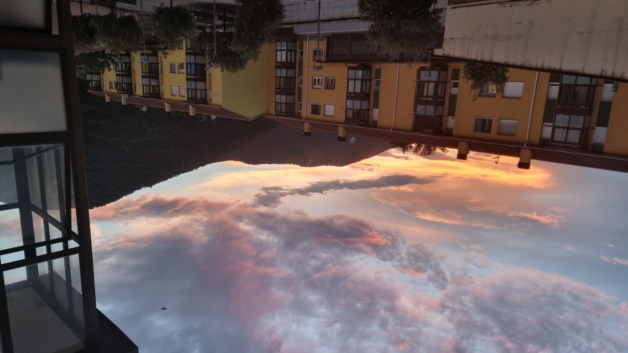 The image depicts a sunset over the town of Sant Joan Les Fonts, Spain. In the foreground, a building with yellow facades and dark balconies is visible. The sky above is filled with dramatic clouds, painted in hues of orange, pink, and purple, indicating the late hour of the day. A dark, tree-covered mountain range forms the backdrop, with the silhouette of the town's buildings in front of it. A bird is seen flying in the upper right portion of the sky. The scene is captured from an elevated perspective, possibly a balcony or a higher floor of another building, as indicated by the partial view of a window frame on the right side of the image.