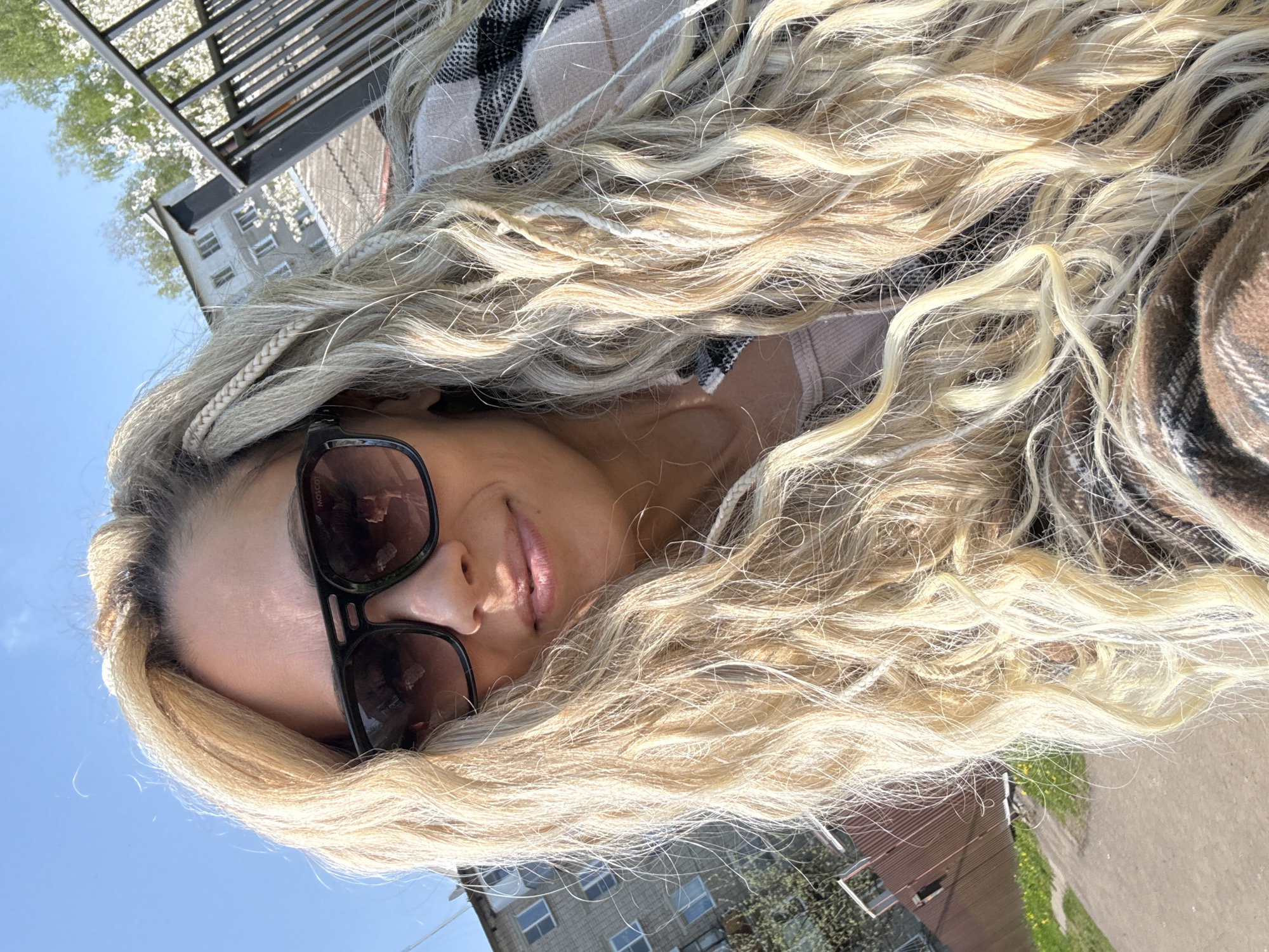 A fair-skinned woman wearing sunglasses is featured in a close-up shot outdoors in Rusanovo, Russia. She is lying down, and her long, wavy blonde hair is spread out around her face and shoulders. She is wearing a beige plaid scarf or blanket and a light-colored top. The background shows a blue sky, a building with windows, some greenery, and a metal railing or structure.