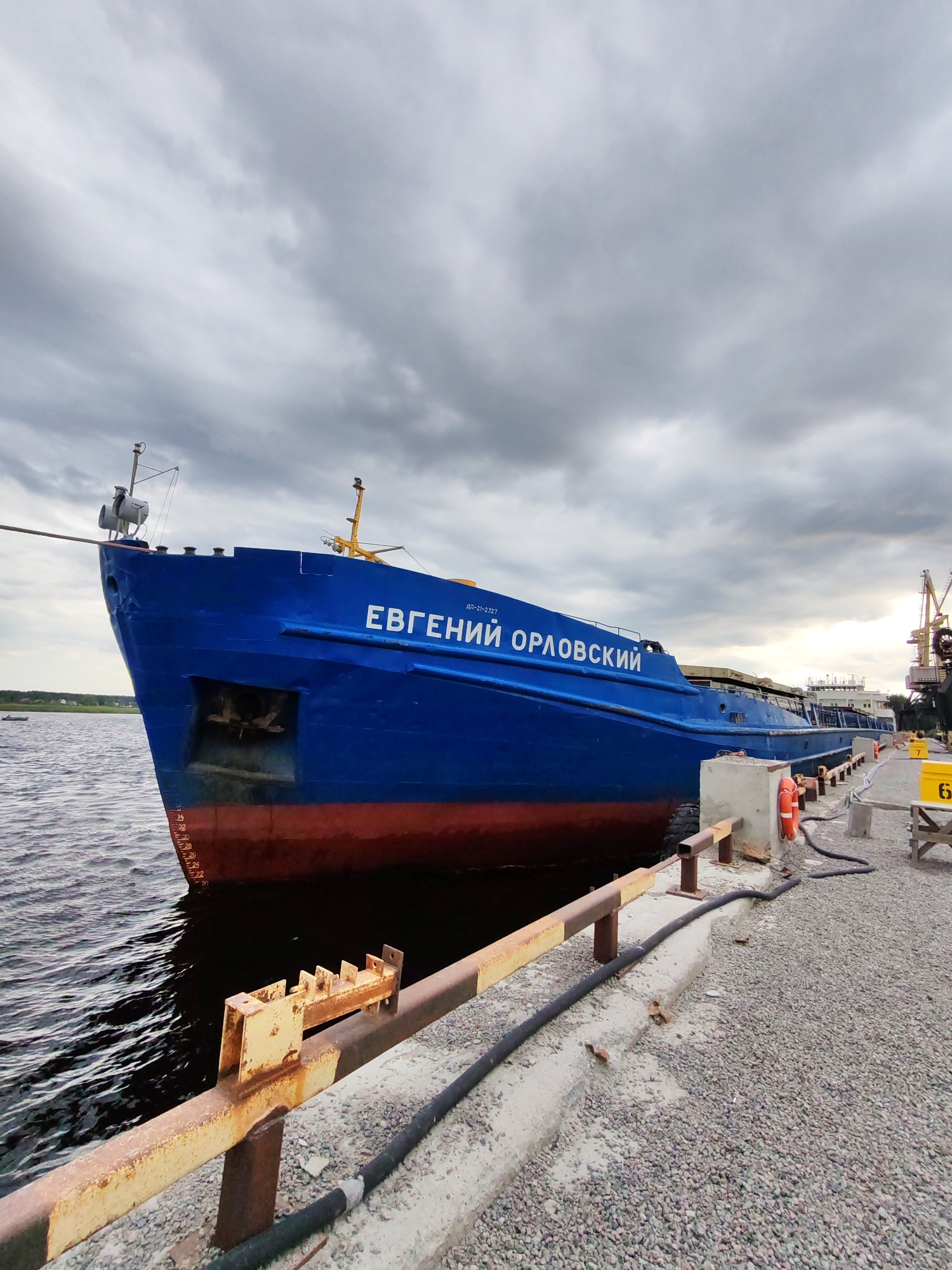 A bright blue cargo ship, named "ЕВГЕНИЙ ОРЛОВСКИЙ," is docked at a port under a cloudy sky. The ship is positioned diagonally in the frame, with its bow facing the viewer's left. The hull of the ship is painted a deep blue with a red waterline. The vessel is moored to a concrete pier, which is covered in gravel and has yellow and brown railings. Various industrial structures, including cranes and yellow containers, are visible in the background to the right. The water next to the ship appears dark and rippled. A black hose runs along the pier. There are no people visible in the immediate vicinity of the ship or the pier. The overall impression is one of an industrial port scene. The location is the city of Shal'skii, Russia.