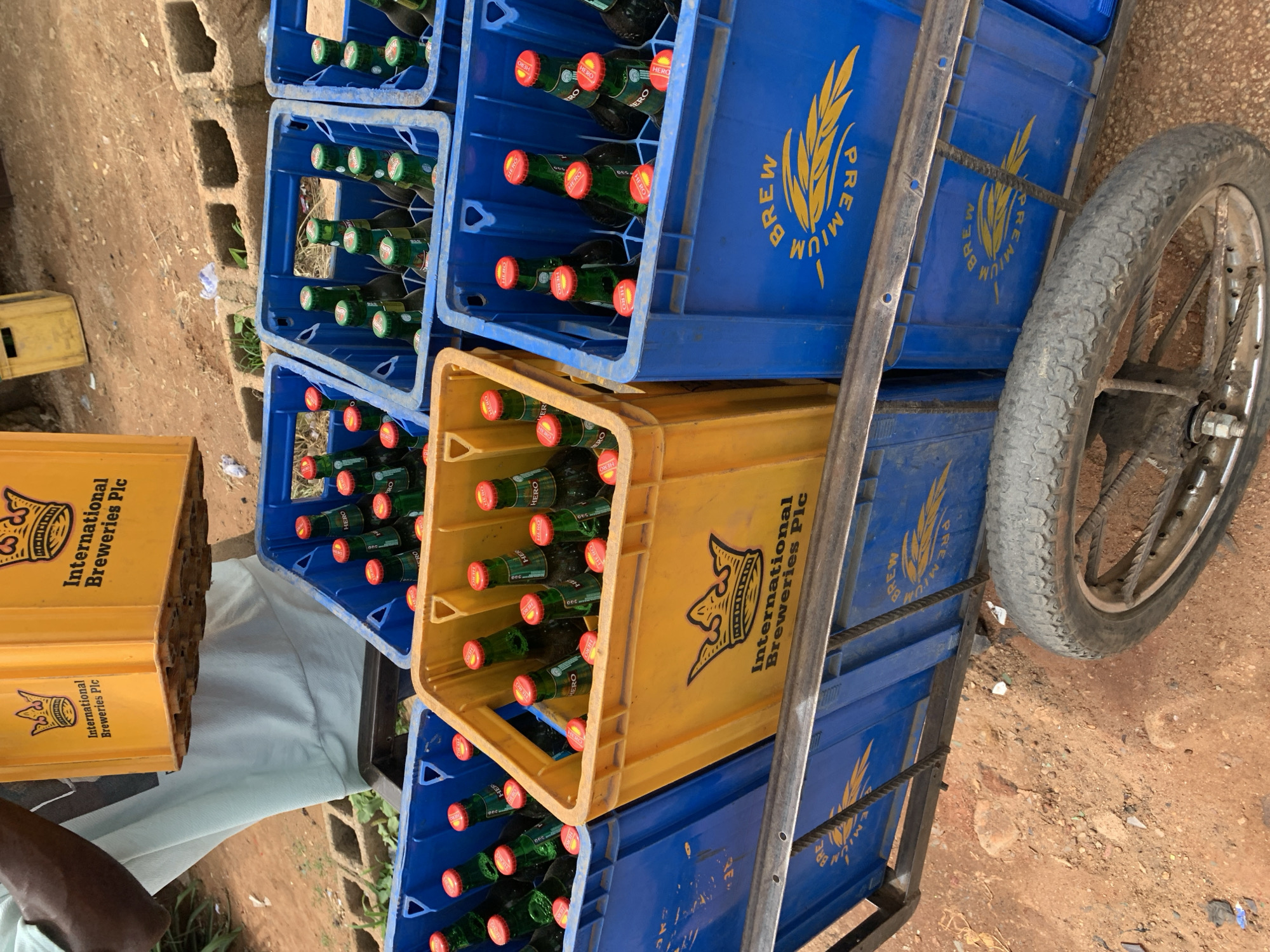 This image captures a street scene in New Stadium, Nigeria, detailing the commercial transport of bottled beverages on a manual trolley. A person, identified by a visible dark-skinned arm and light-colored t-shirt in the upper left, is handling a yellow plastic crate. This crate, and an identical one positioned on the trolley, bears a crown logo and the text "International Breweries Plc." Both yellow crates contain green glass bottles topped with red caps, many of which are marked with "HERO." Multiple blue plastic crates are also loaded onto the metal-framed trolley. These blue crates similarly hold green glass bottles with "HERO" branded red caps. The blue crates are adorned with a stylized wheat sheaf logo and the text "PREMIUM BREW." The trolley's single visible wheel, equipped with a dark, treaded tire and metal spokes, is prominent in the lower right. The ground surface is unpaved reddish-brown dirt, with concrete blocks and minor debris visible in the background.