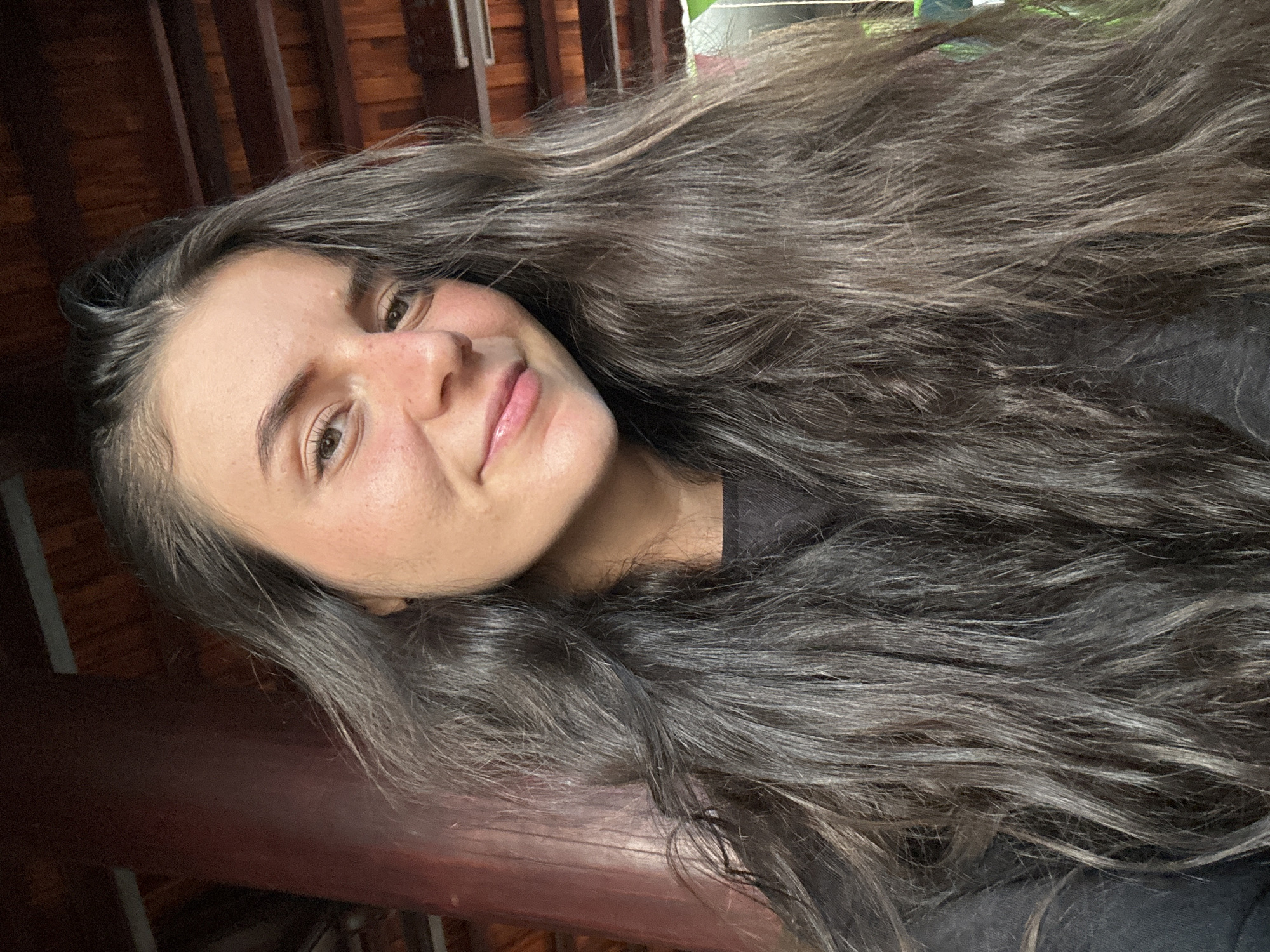 A woman with long, dark hair is pictured in a close-up, angled portrait shot. Her face is turned slightly to the side, with a gentle smile. The background appears to be a wooden structure, possibly part of a building or veranda. The photograph may have been taken in an outdoor or semi-outdoor setting. There is no visible information to confirm if the photo was taken in the city of Mũi Né, Vietnam.