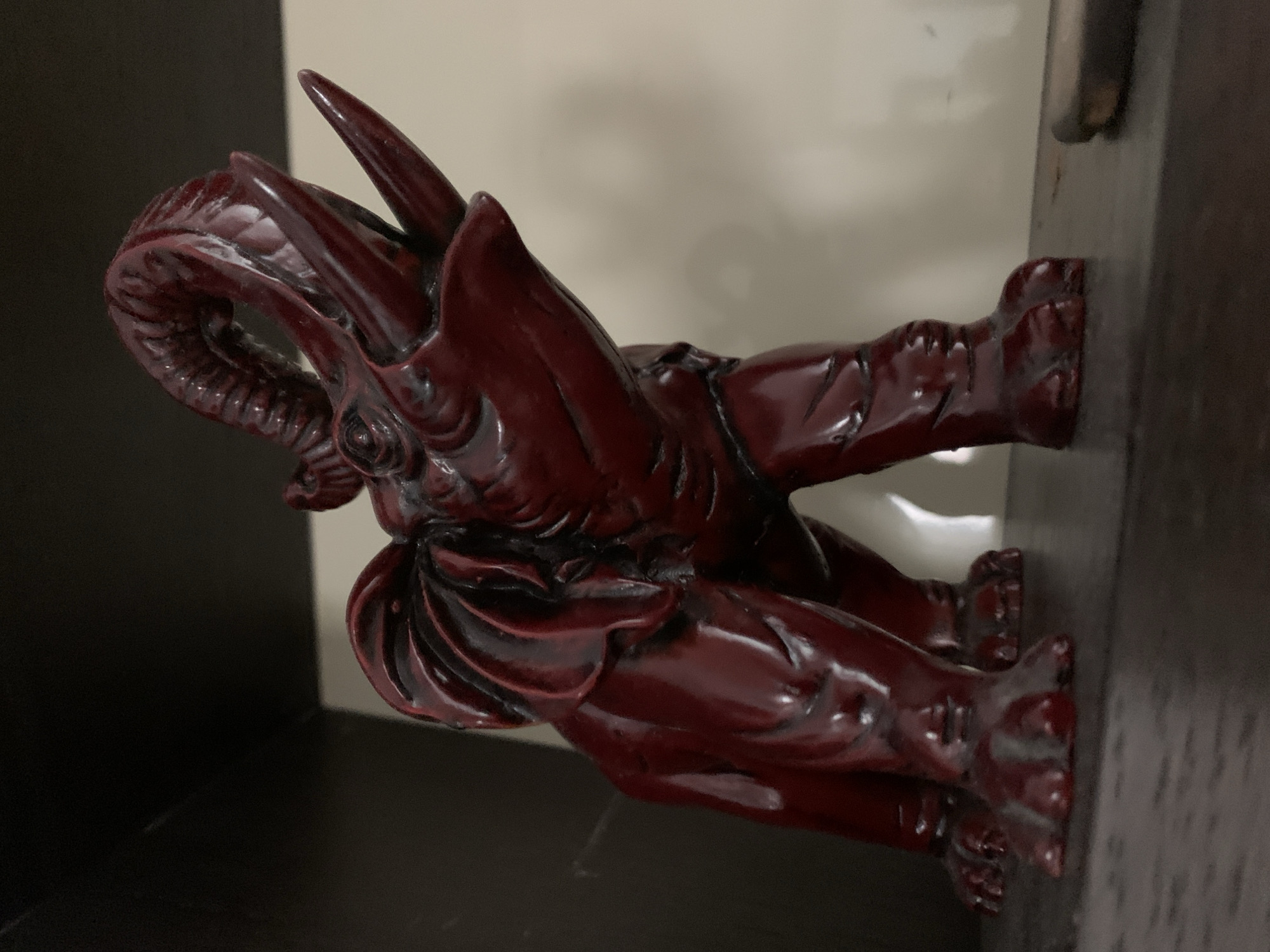 A maroon-colored statuette of a dragon-like creature is positioned on a dark shelf against a beige wall. The statuette is oriented horizontally in the image, with its head on the left and feet on the right. The details of the statuette are distinct, showing the scales, horns, and intricate detailing of the creature. The setting appears to be indoors, with a basic shelf and wall construction. There is no clear evidence to directly link the image to the Vozdovac Urban Municipality, Serbia.