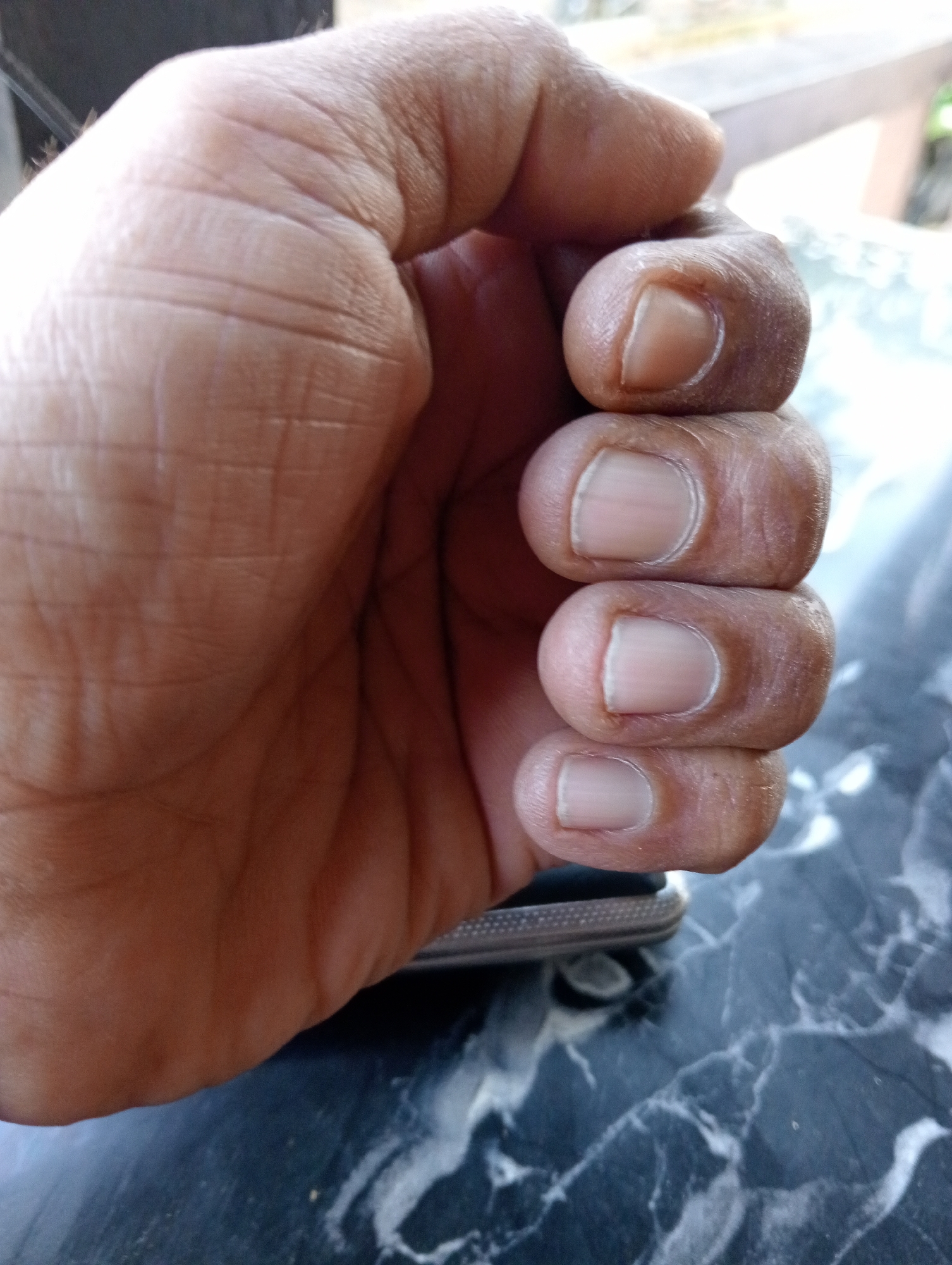 A close-up image shows a person's left hand resting on a dark, mottled surface, possibly a marble or granite table. The hand is palm-down, fingers stacked tightly together. The skin is light brown with visible creases and wrinkles. Fingernails are short, rounded, and appear clean. A small portion of a metallic object, possibly a smartphone, is visible under the hand. The background is slightly blurred, but a wooden structure can be discerned. The photo was likely taken outdoors in daylight. The city of Lhokseumawe, Indonesia is not discernable.