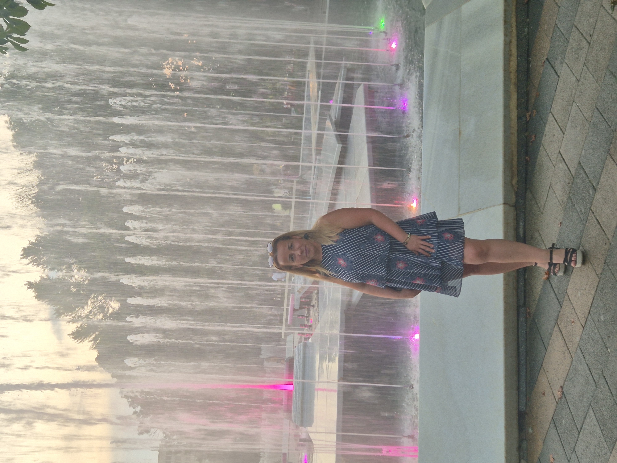 A medium shot captures a woman standing in front of a fountain at dusk. The woman, with blonde hair and wearing a striped dress and sandals, poses with her hands on her hips. The fountain, illuminated by pink and green lights, sprays water into the air. The background shows trees and buildings in the distance. The ground in the foreground is paved with interlocking stones.