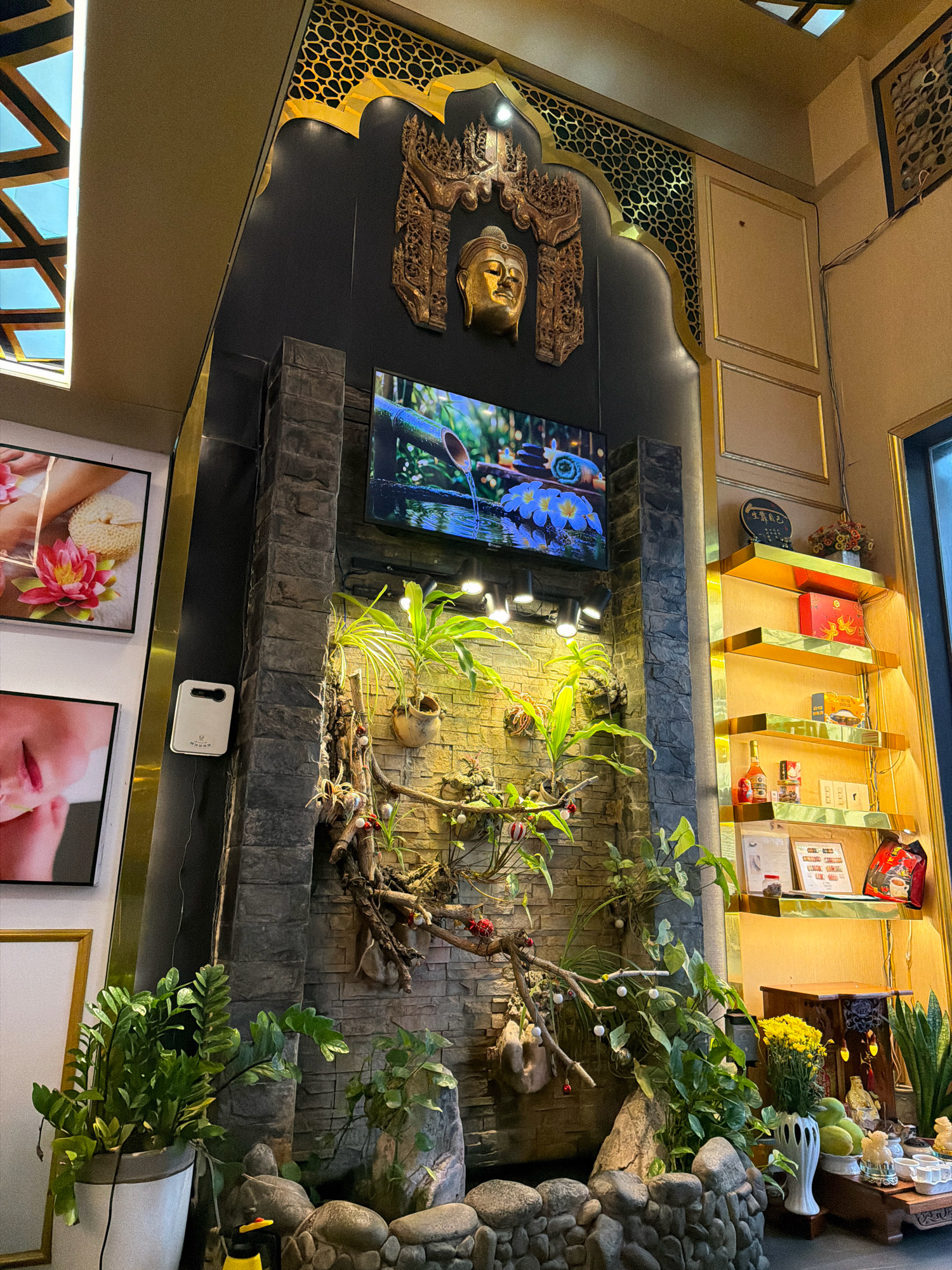 The image depicts the interior of a spa or wellness center in Nha Trang, Vietnam. A large, ornate golden archway with intricate latticework frames a central wall feature. This feature is a multi-layered stone structure with a small television screen displaying a nature scene, and a decorative golden Buddha head mounted above it. Lush green plants are integrated into the stonework, with some hanging from branches and others in pots. To the left of this central feature, there are framed pictures on the wall, one of which shows hands performing a spa treatment with a lotus flower. To the right, golden shelves are illuminated, displaying various products, boxes, and decorative items. In the foreground, at the bottom of the stone structure, there is a small water feature surrounded by stones and more greenery. The overall ambiance is serene and decorated with an Asian aesthetic, common in spas.
