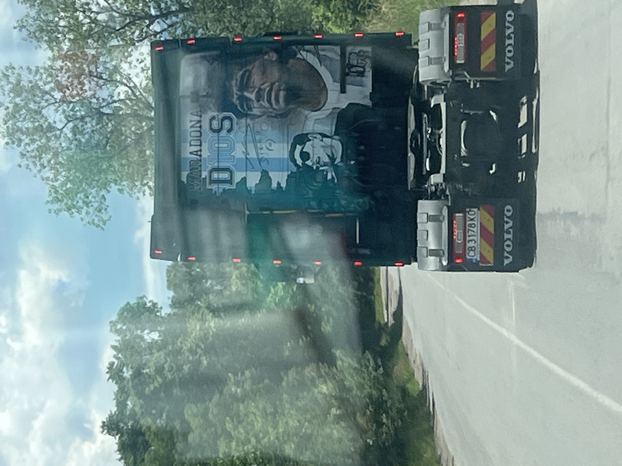 The image shows the rear of a Volvo truck driving on a road. The truck's trailer is adorned with a mural of Diego Maradona. The mural features two portraits of Maradona, one larger and more prominent, and a smaller one wearing sunglasses. The word "MARADONA" is written above "DIOS," which translates to "God." The truck has a license plate that reads "CB 3178 KO." The truck is on a paved road that appears to be in a rural or semi-rural area, with trees lining the sides of the road. The sky is partly cloudy.