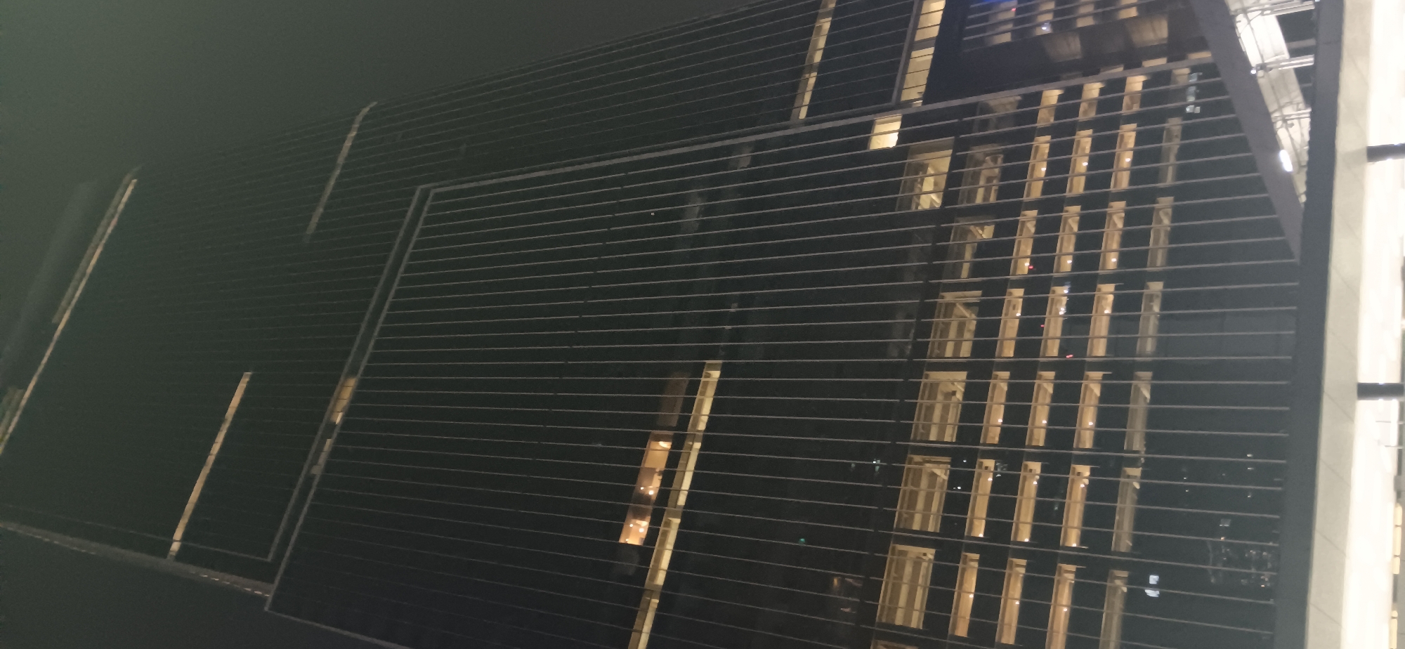 A low-angle, full shot captures the exterior of a modern, glass-faced skyscraper at night. The building dominates the frame, its dark facade punctuated by faint horizontal bands of light at various levels, suggesting interior illumination. Vertical lines from the building's structure crisscross the image, creating a sense of depth and scale. The sky above is dark and featureless, indicating nighttime conditions. The skyscraper's reflective surface mirrors the faint lights, creating a slightly distorted yet elegant effect. The composition emphasizes the building's height and sleek, contemporary design against the dark urban backdrop. The image does not contain any identifiable people or specific landmarks that would indicate its location in Central Jakarta, Indonesia.