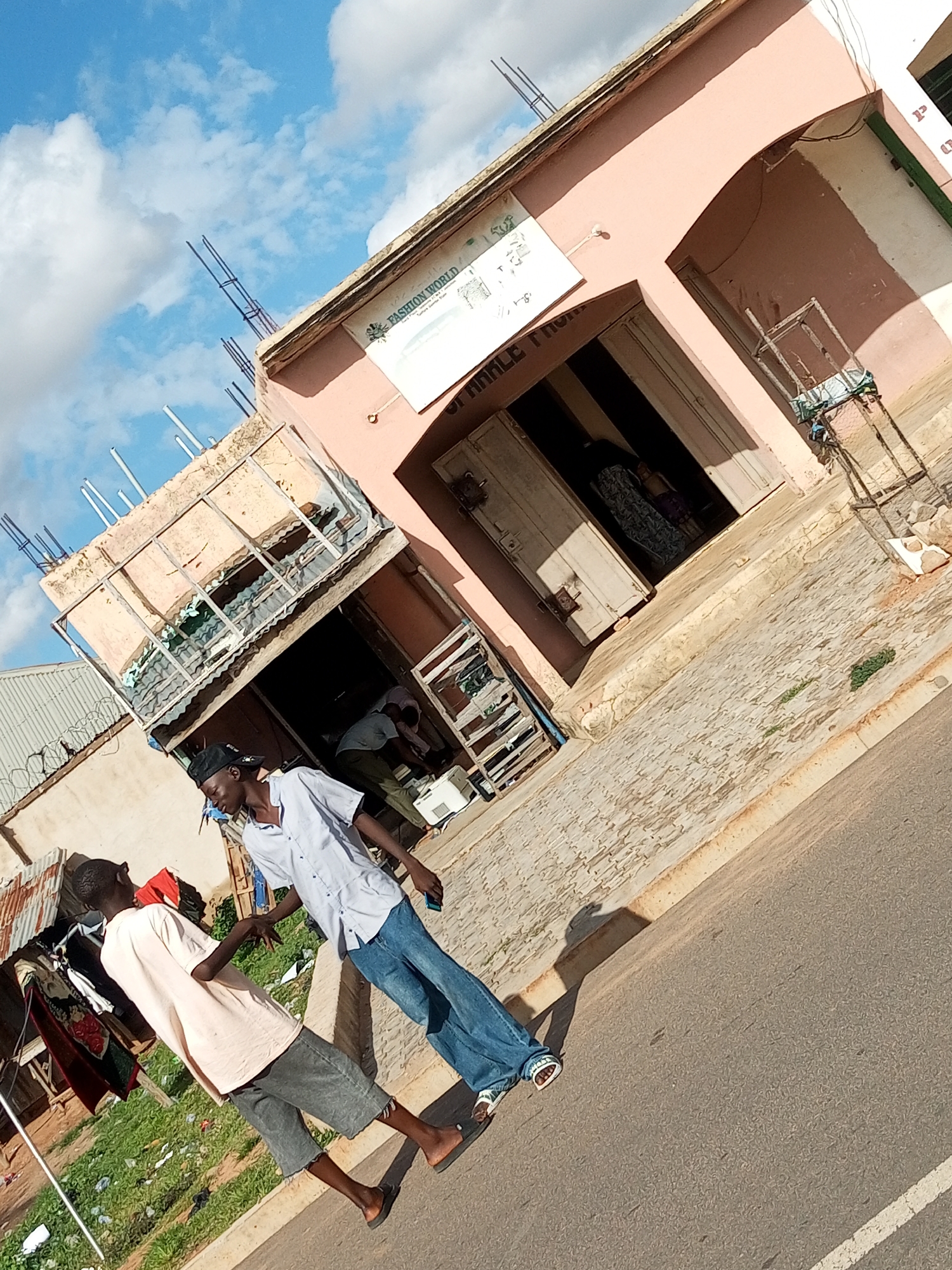 In an outdoor, urban setting, two young men converse on a street in front of a pink building with an arched entryway and a store displaying signage that reads "FASHION WORLD." The man on the left wears a light pink t-shirt and grey shorts, while the man on the right sports a light blue shirt, blue jeans, and a black baseball cap. They stand on an asphalt road with a paved walkway adjacent to it. To the left of the men, clothing is displayed on a rack, suggesting a market or retail area. The background shows various structures, including a corrugated metal roof, and the sky is filled with white clouds against a blue backdrop. The image is taken from a slightly elevated angle and is rotated clockwise.