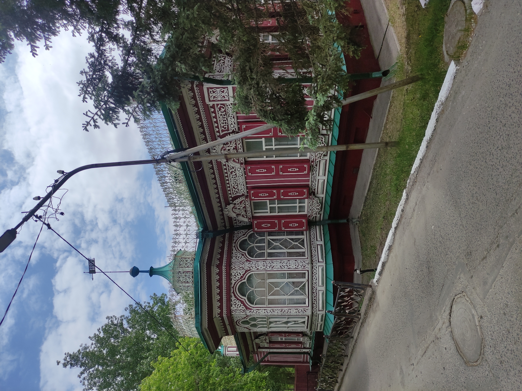The image shows a wooden, ornate house with intricate carvings in Pavlodar, Kazakhstan. The house is painted in red, white, and green. The facade has large windows and decorative shutters. The house appears to be located on a street with trees nearby. A light pole is visible in front of the house. The sky is blue with some clouds. A sidewalk and street are in the foreground. There are no people in the image. A manhole cover is visible in the street.