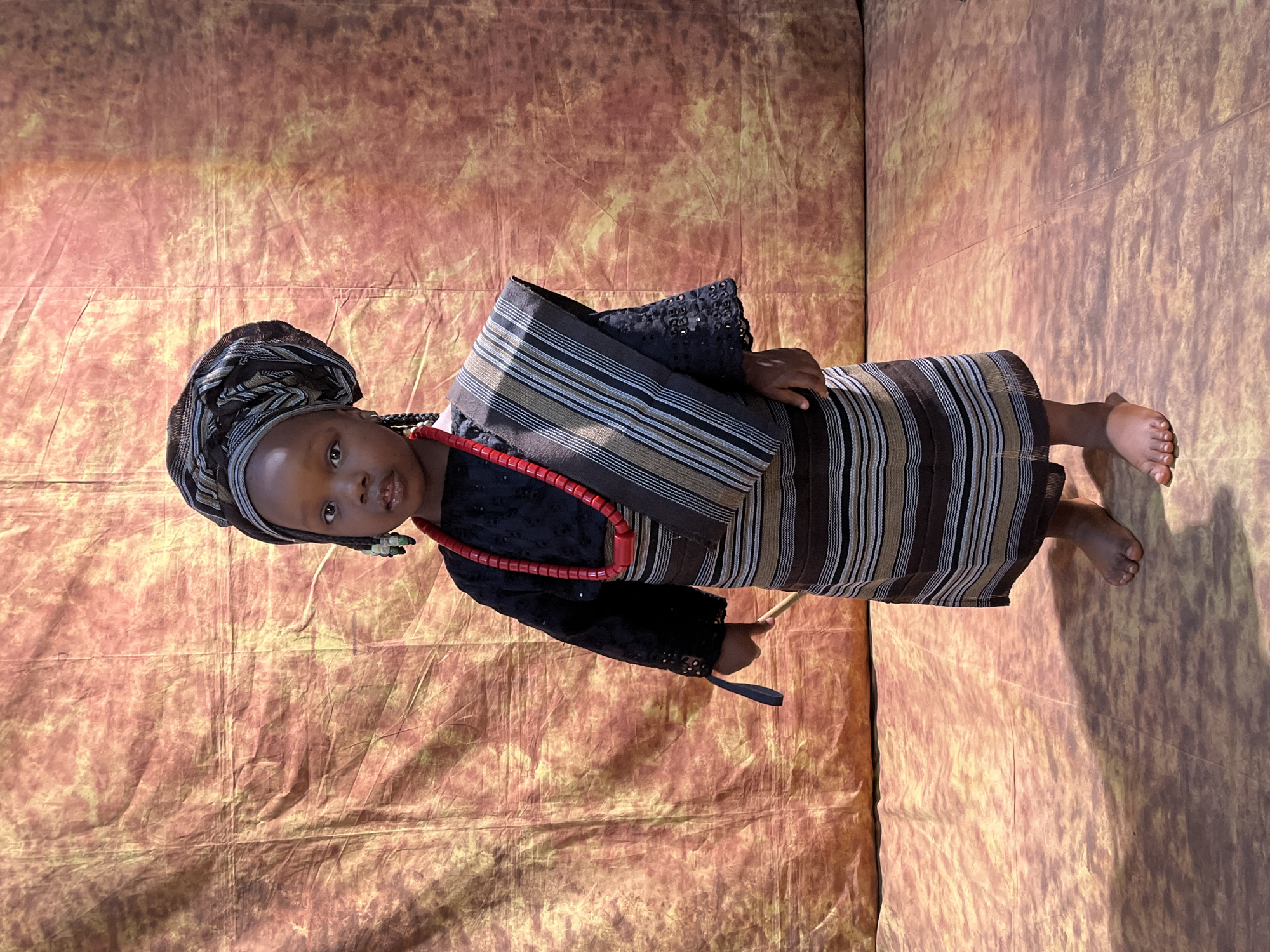 A young Black child, likely under five years old, stands facing the camera in a studio setting. The child is dressed in traditional Nigerian attire, including a patterned headwrap, a black lace top with detailed sleeves, a layered beaded necklace in red, and a striped skirt. A patterned sash is draped over the child's left shoulder. The child is holding a thin, light-colored stick or rod in their right hand. The background consists of a textured, mottled fabric in shades of brown, orange, and yellow, resembling a rustic or earthy backdrop. The floor is also textured with a similar color palette. The lighting appears to be artificial, creating shadows on the floor. The image's composition suggests a formal portrait, possibly for cultural documentation or a special occasion. The child's expression is neutral. No specific location in Jos, Nigeria, can be identified from the visual information provided.