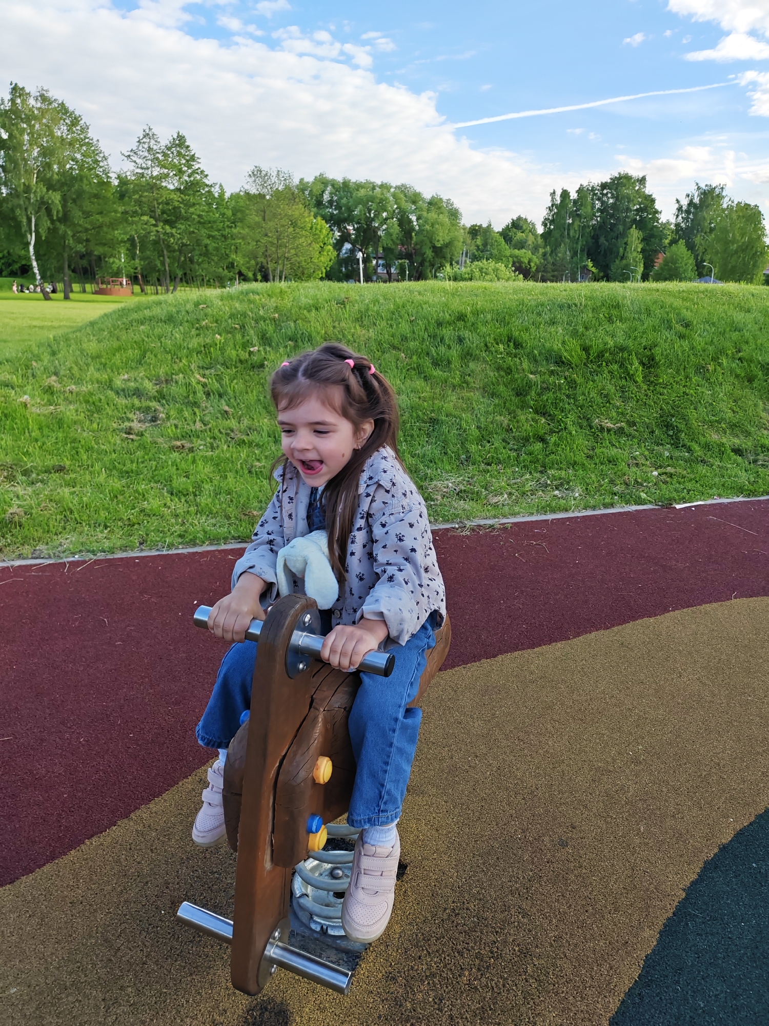 A young girl is riding a wooden rocking horse in a park. She is wearing a light patterned jacket, blue jeans, and pink sneakers. Her hair is styled in pigtails with pink ties. She is smiling broadly. The rocking horse is made of wood and metal, with yellow and blue details. The ground is covered in a soft, multi-colored material. In the background, there is a grassy hill and trees. The sky is blue with white clouds. The location is Bronnitsy, Russia.