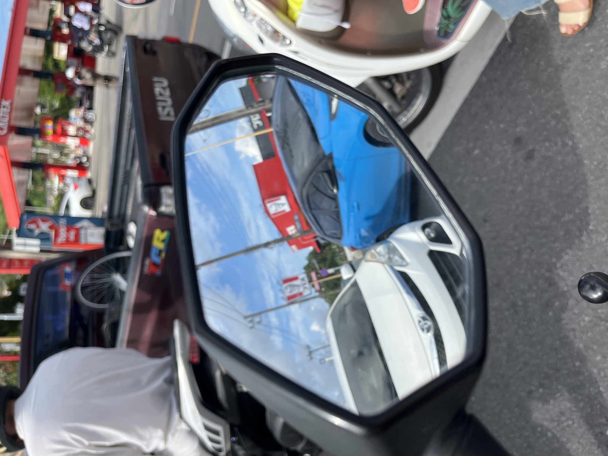 The image is a rearview mirror reflection of a street scene in Phuket City Municipality, Thailand. In the reflection, a blue car is stopped in traffic next to a white car. Behind them, a red KFC restaurant building is visible. The sky is blue with scattered clouds. On the right side of the reflection, a white scooter is parked. In the foreground of the image, a person is sitting on a motorcycle, facing away from the camera. Part of a red truck with "ISUZU" branding is also visible in the scene. The overall environment appears to be an urban street with commercial buildings and ongoing traffic.