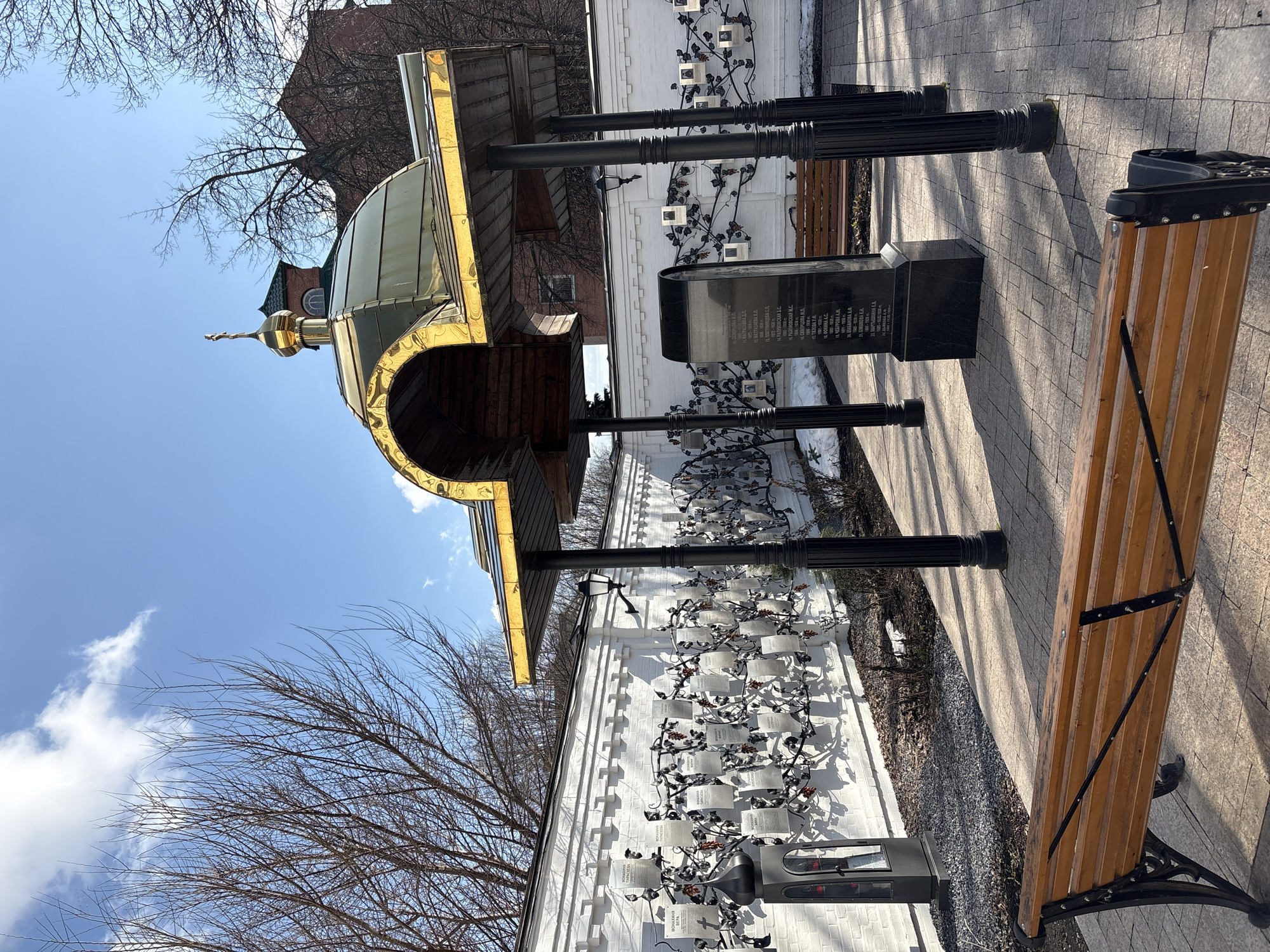The image presents a scene in what appears to be a public space in Ulyanovsk, Russia, featuring architectural elements and memorials. The most prominent structure is a small, ornate building with a golden dome topped by a cross, indicative of Russian Orthodox architecture. Adjacent to the building is a white brick wall adorned with dark metal decorative elements resembling vines, and small, square plaques attached to these vines. A dark stone monument stands nearby, inscribed with text in Russian. A park bench with wooden slats and black metal supports sits to the right of the monument. The ground is paved with light-colored stones, and a bare tree is visible in the background against a blue sky with scattered clouds. The text in the image is partially readable, with surnames like "MATRAZAEVA", "RAZUMOVSKAYA", "FOKINA", "CHEBUROVA", "SHISHKOVA", and "ARTYSHKINA" identified.