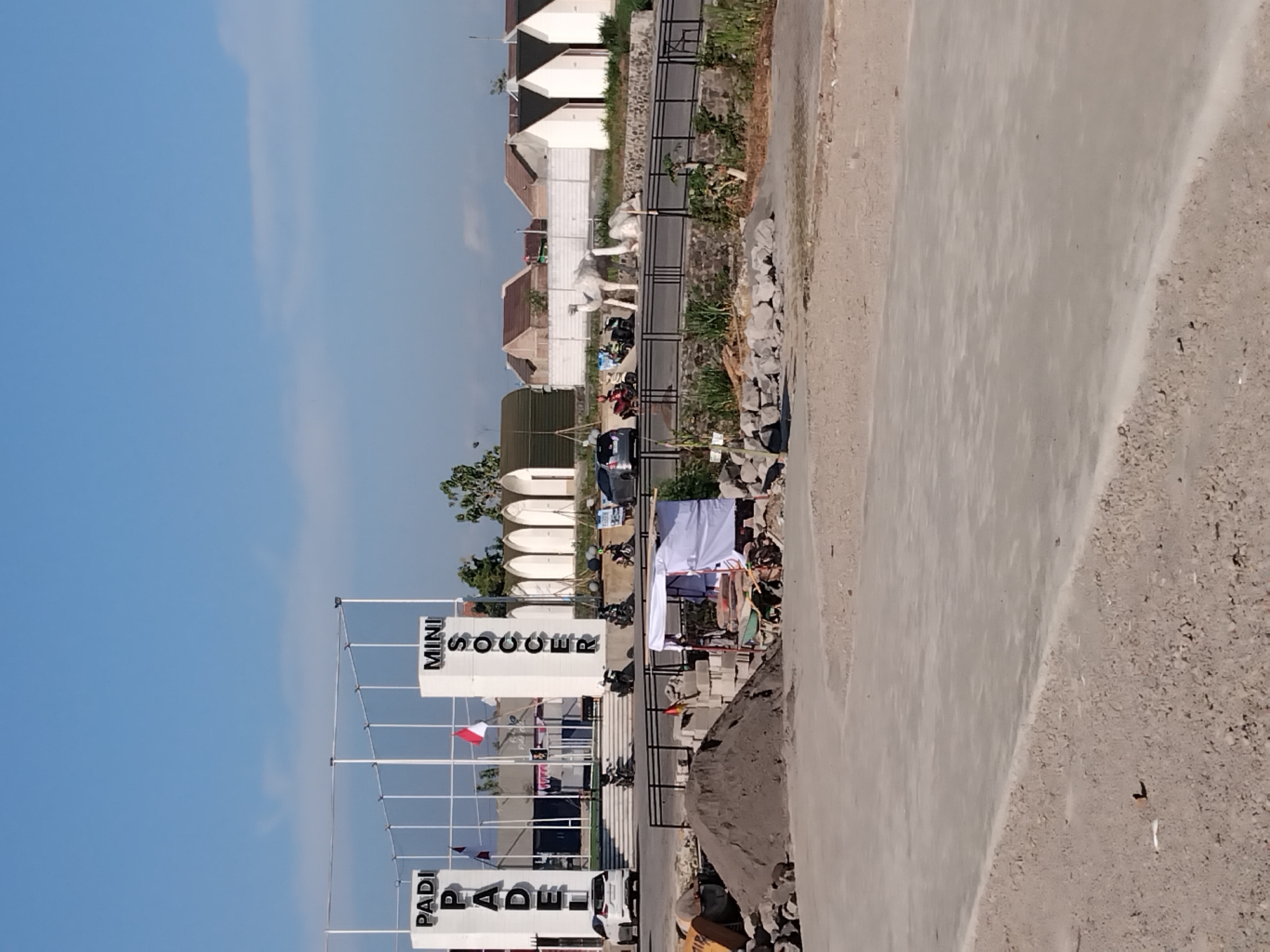 In the city of Papahan, Indonesia, an outdoor recreational facility features a "PADI PADEl" court and a "MINI SOCCER" field. Two white pillars with the respective names are visible. In the foreground, there is a construction site with piles of sand and stones, along with a white tent. Further in the background, several houses with white walls and dark roofs are arranged in a row. Two camel statues are placed near a stone wall, and various vehicles, including motorcycles and cars, are parked around the facility. The sky is clear and blue.