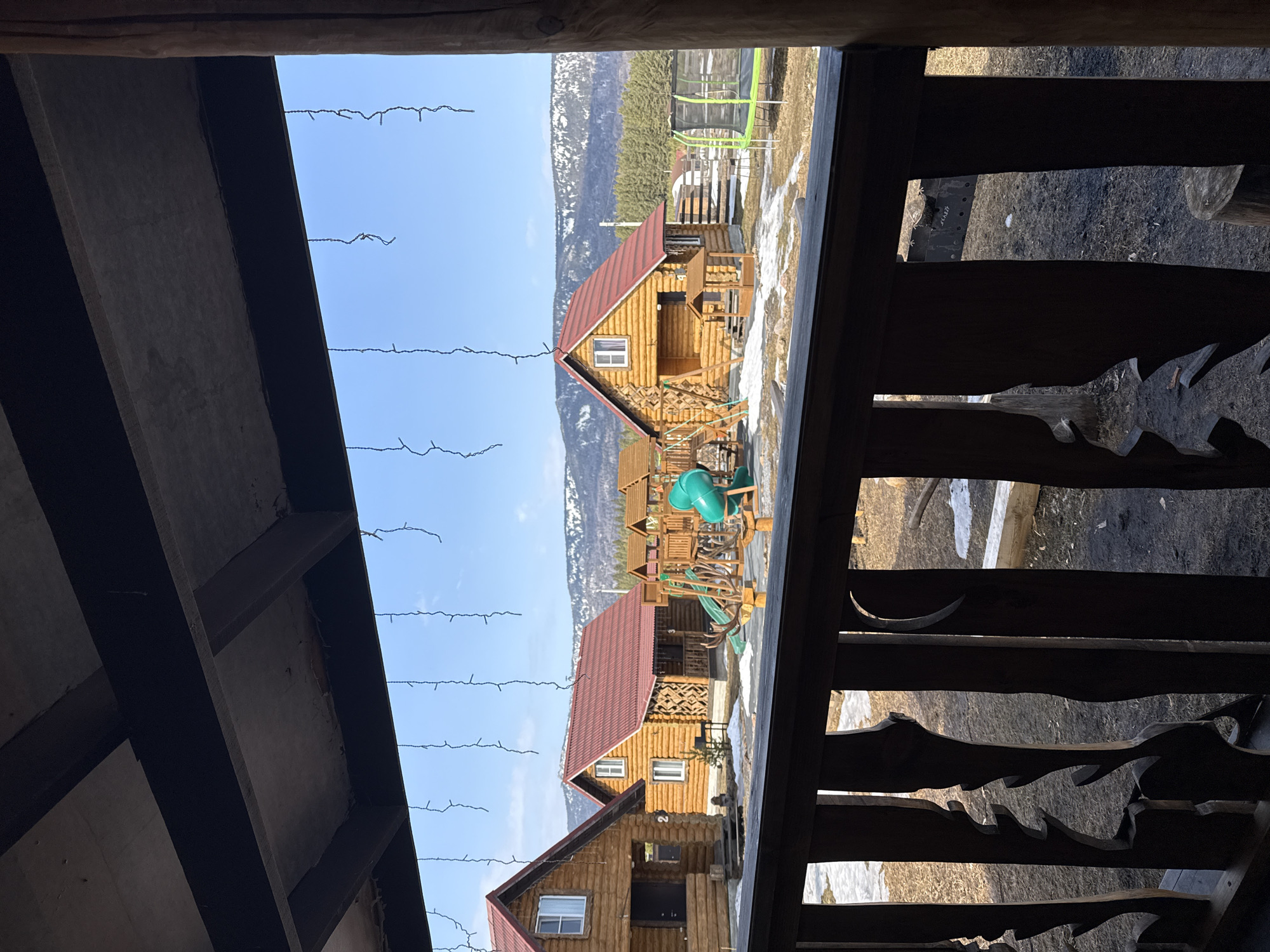 The image displays an outdoor scene in Tyulyuk, Russia, captured from beneath a dark wooden overhang and through a decorative wooden railing with carved balusters. Strands of white string lights are suspended from the overhead structure. The midground features a recreational area comprising several log cabins. Three prominent log cabins, constructed from light-toned logs with reddish-orange corrugated metal roofs, are visible. The leftmost cabin has a dark door, white-framed windows, and the number "2" affixed to its side. The central cabin, also with white-framed windows, displays the number "3" on a porch post and features decorative wooden lattice. A wooden children's playground structure, including two green slides, swings, and carved antler-like wooden embellishments, is situated between the cabins. The ground is partially covered with melting snow and exposed dark earth. A green and black trampoline is positioned on the far right. The background reveals a dense line of evergreen trees, beyond which a range of snow-dusted mountains rises under a clear blue sky. No individuals are present. No actions or interactions are depicted.