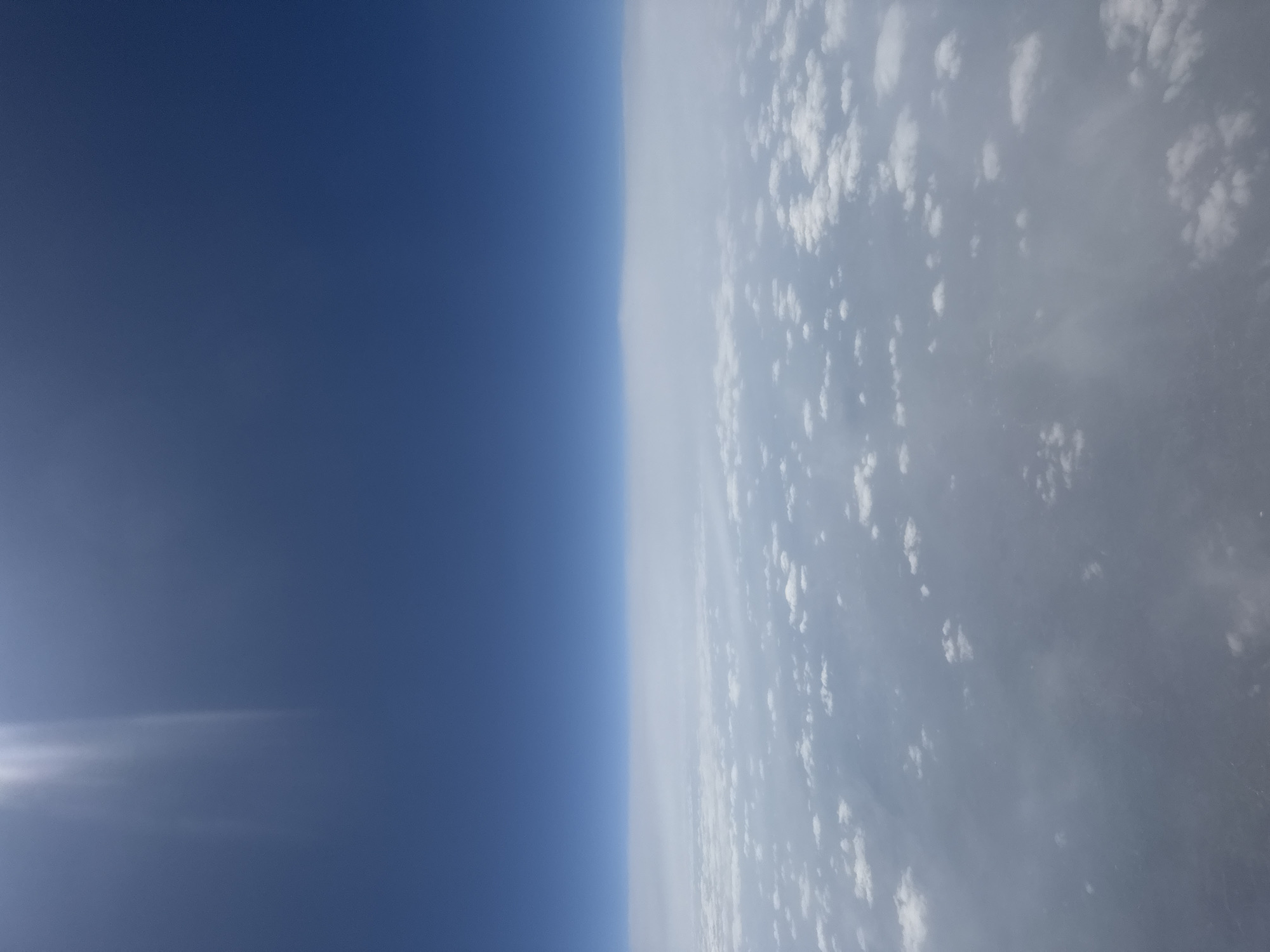 This image presents an expansive aerial view, likely captured from a high altitude such as an aircraft or near-space, looking across the Earth's atmosphere. The scene is distinctly divided into two primary sections by a clear, vertical horizon line (when viewed with the blue expanse on the left and clouds on the right).
The left portion of the image is dominated by a vast, deep blue sky, indicative of the upper atmosphere or space. The color transitions subtly, becoming a slightly lighter blue closer to the horizon line and deepening to an indigo hue further away. A bright, intense glare, possibly from the sun or a lens flare, is visible in the upper-left quadrant of this blue expanse, highlighting the bright daylight conditions.
The right side of the image features an extensive cloud layer. Numerous white, puffy clouds of varying sizes are scattered across a broader, greyish-blue atmospheric haze, creating a textured and layered appearance. Some areas show denser concentrations of these cumulus-like clouds, while others are more sparse, allowing the underlying atmospheric haze to be more prominent. Faint rays of light or atmospheric scattering effects can be observed within the cloud formations.
There are no people, man-made structures, or identifiable landforms visible in the image. The focus is entirely on the atmospheric elements and the horizon. The bright illumination confirms it is daytime. No text is present. While the image itself does not offer direct cues, the provided context suggests this view is over or near Sofia, Bulgaria, offering a unique perspective of its skies from above.