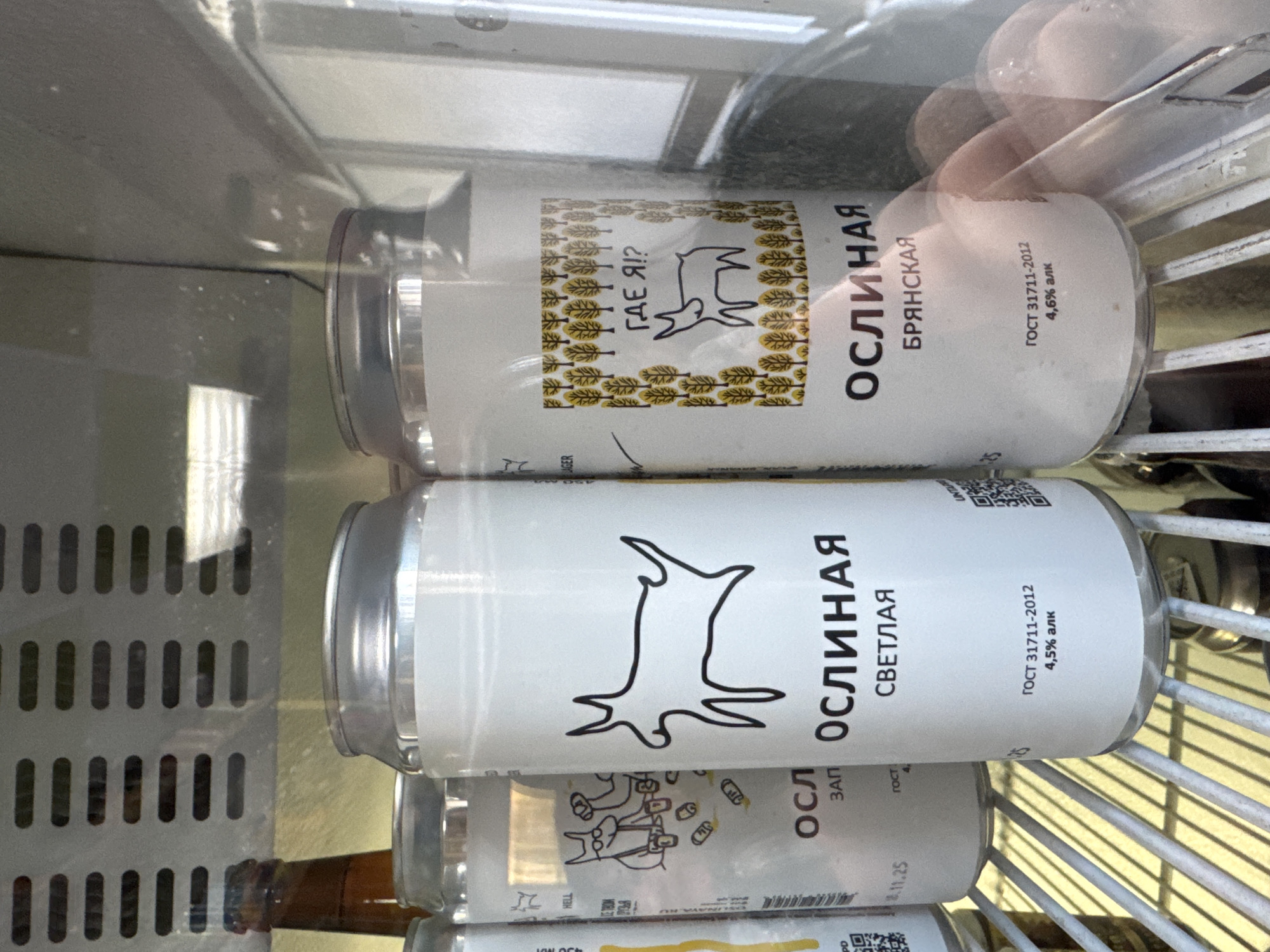 The image shows several cans of "Oslinaya" beer in a refrigerated display case, possibly in a store in an unspecified city. The cans have white labels featuring a stylized donkey graphic. One variety is "Bryanskaya" with 4.6% alcohol, and another is "Svetlaya" with 4.5% alcohol. A brown glass bottle is visible in the lower left corner. A hand is partially visible reaching into the case. The scene suggests a commercial setting.